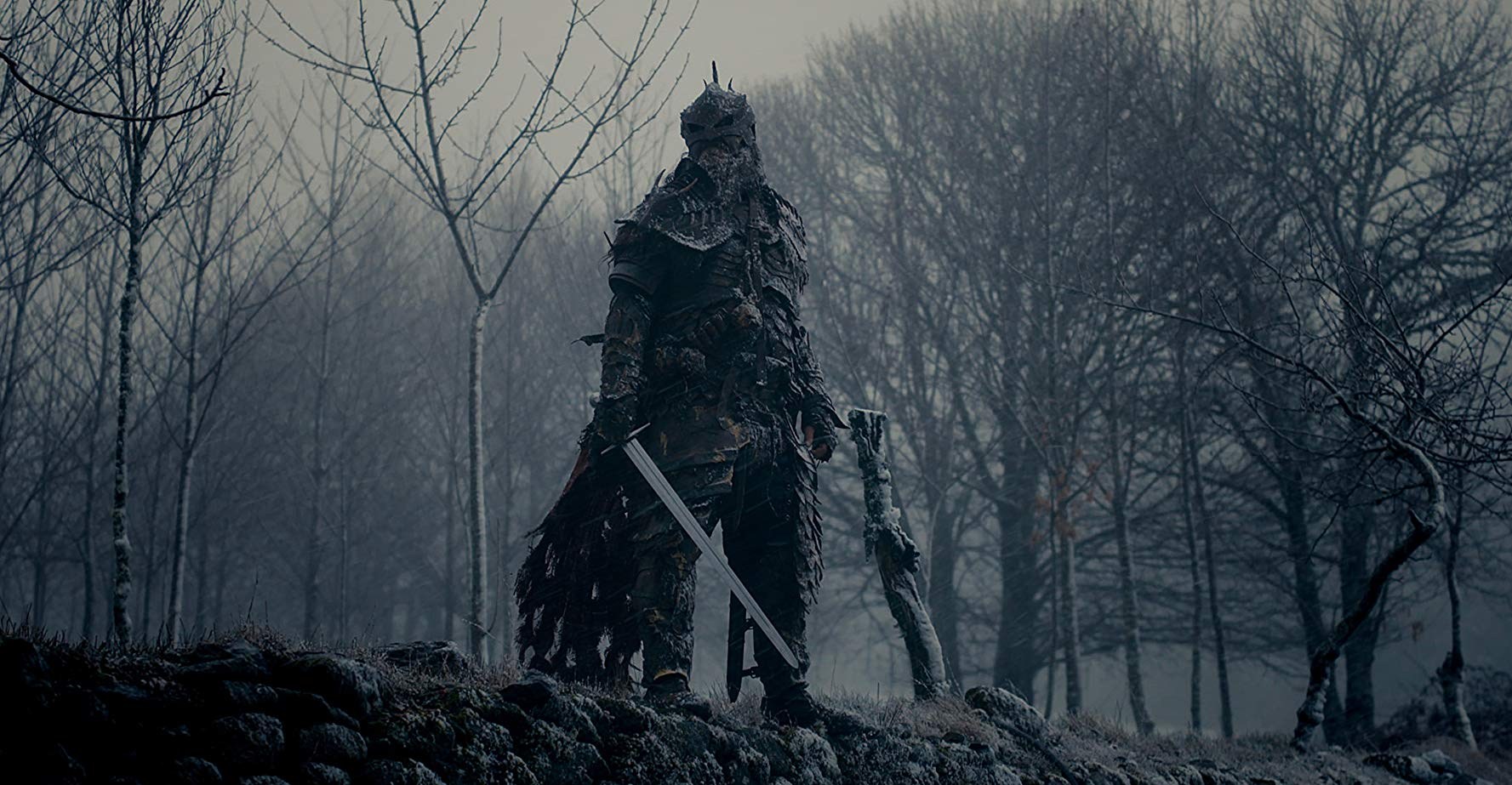 Um guerreiro em armadura preta fica em frente a uma floresta de inverno desolada.