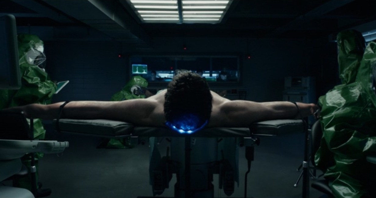 Reed Richards, interpretado por Miles Teller, está amarrado a uma mesa com os braços esticados em uma cena do filme Fantastic Four 2015. 