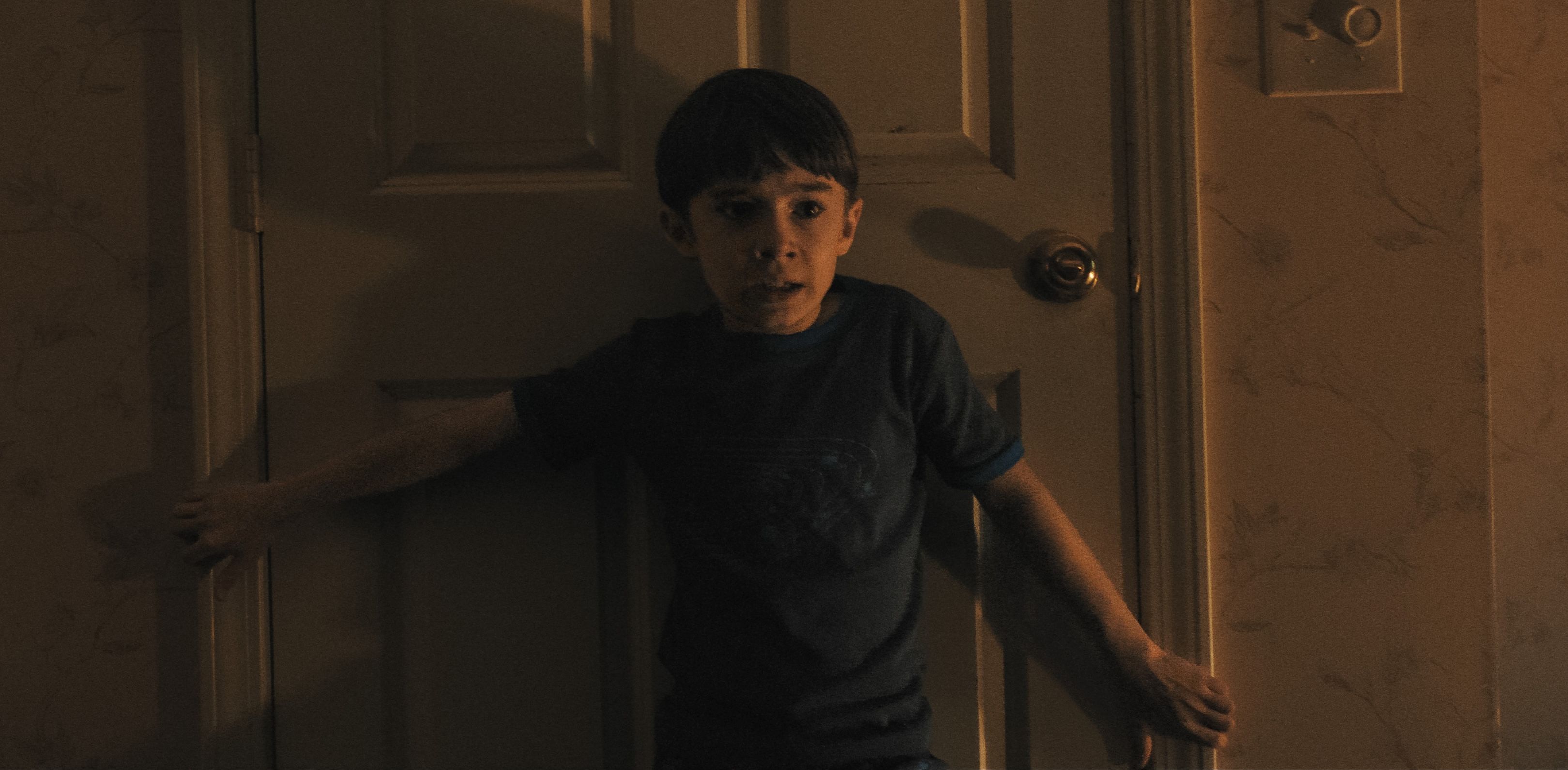 Um jovem menino aterrorizado fica com as costas pressionadas em uma porta fechada e seus braços estendidos para bloqueá -lo em armas