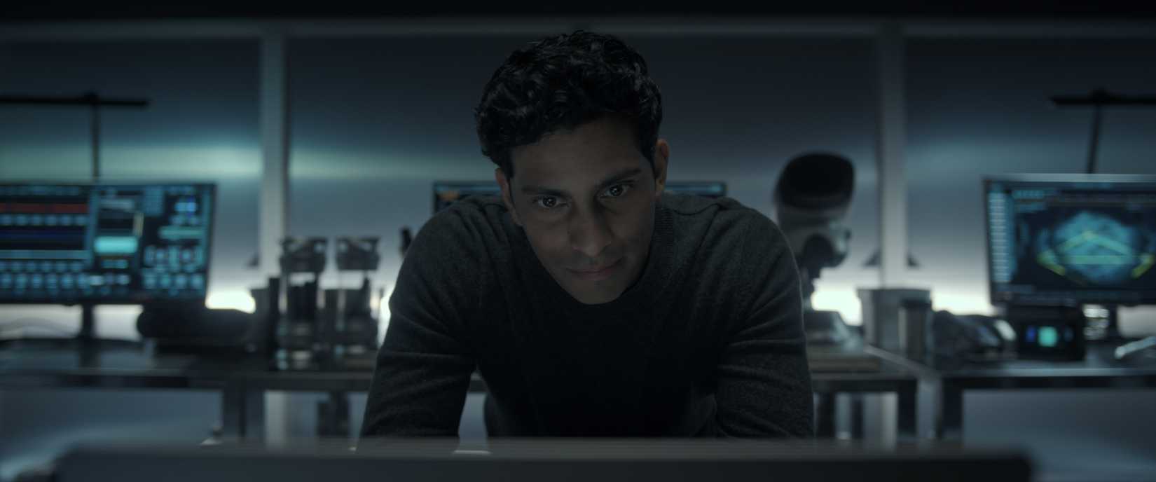 Um homem de frente para a câmera olha para uma tela de computador em um escritório cheio de equipamentos e computadores científicos.