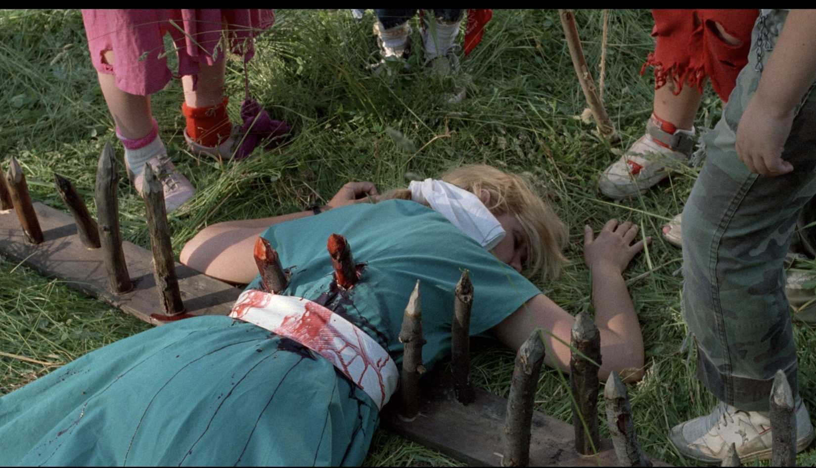 O cadáver de uma mulher, empalado em uma série de picos de madeira curtos saindo de uma prancha, fica na grama em cuidado! Crianças em brincadeira