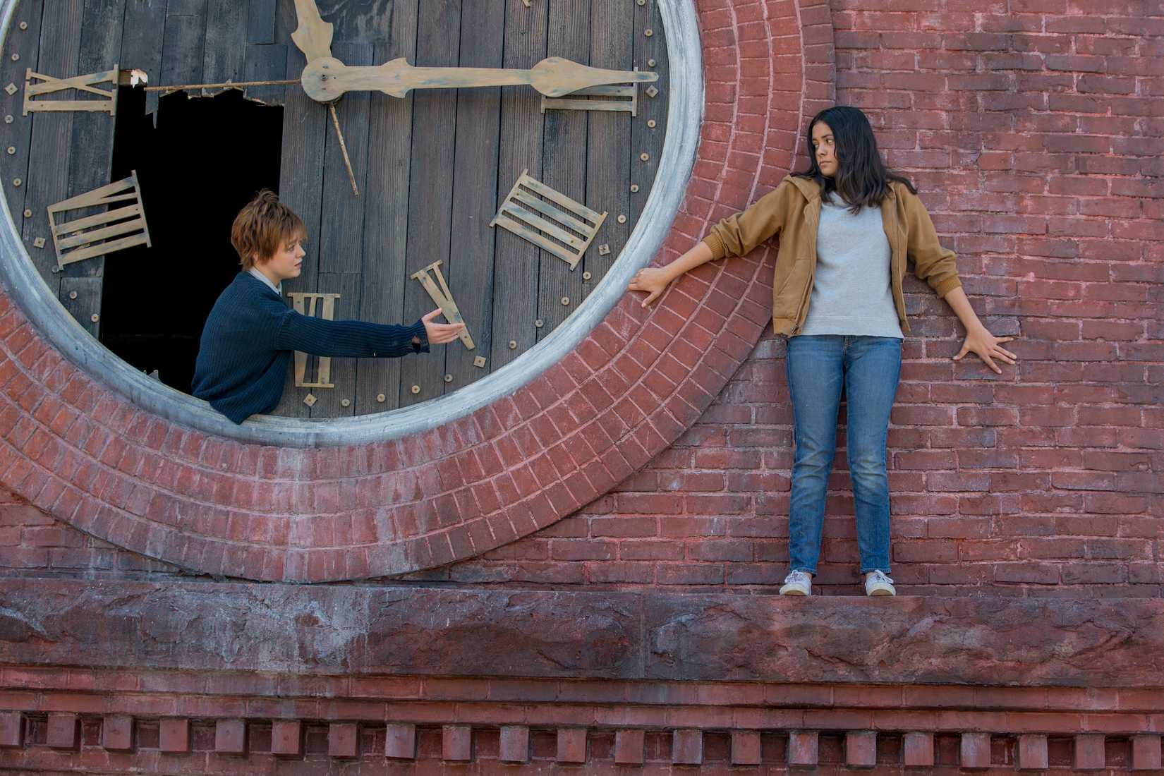 Rahne Sinclair (Maisie Williams) se inclina entre tábuas quebradas em uma face gigante do relógio, estendendo a mão para Dani Moonnstar (Blu Hunt), que está em uma borda na face de tijolos da torre do relógio, pressionou contra a parede, nos novos mutantes do século XX. 