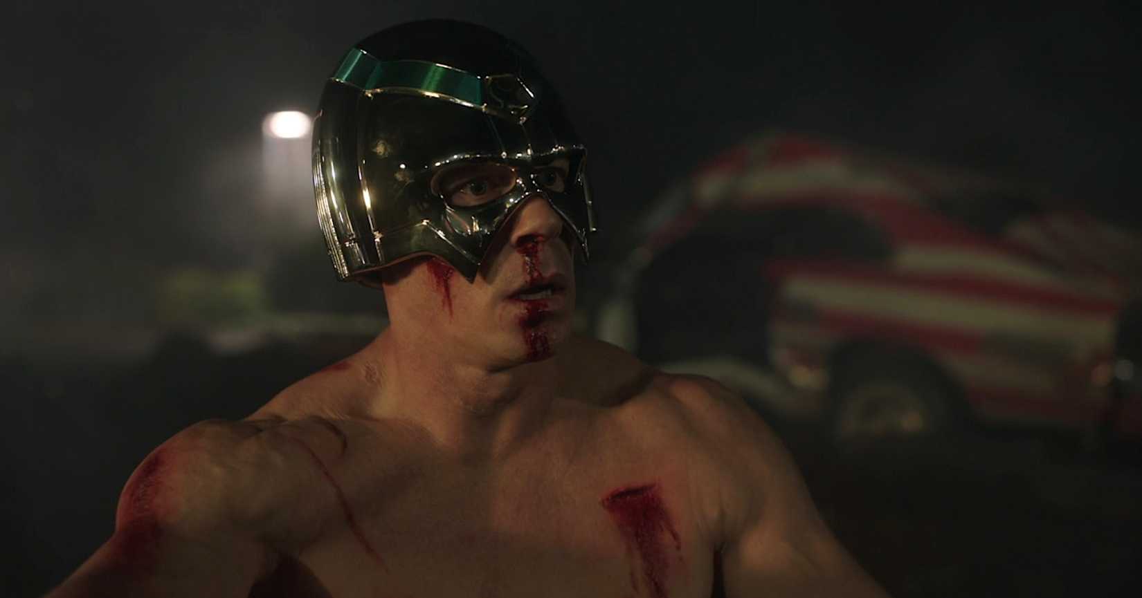 Peacemaker (John Cena), sem camisa, atordoado e coberto de ferimentos sangrando, fica em um estacionamento à noite na 1ª temporada, episódio 1