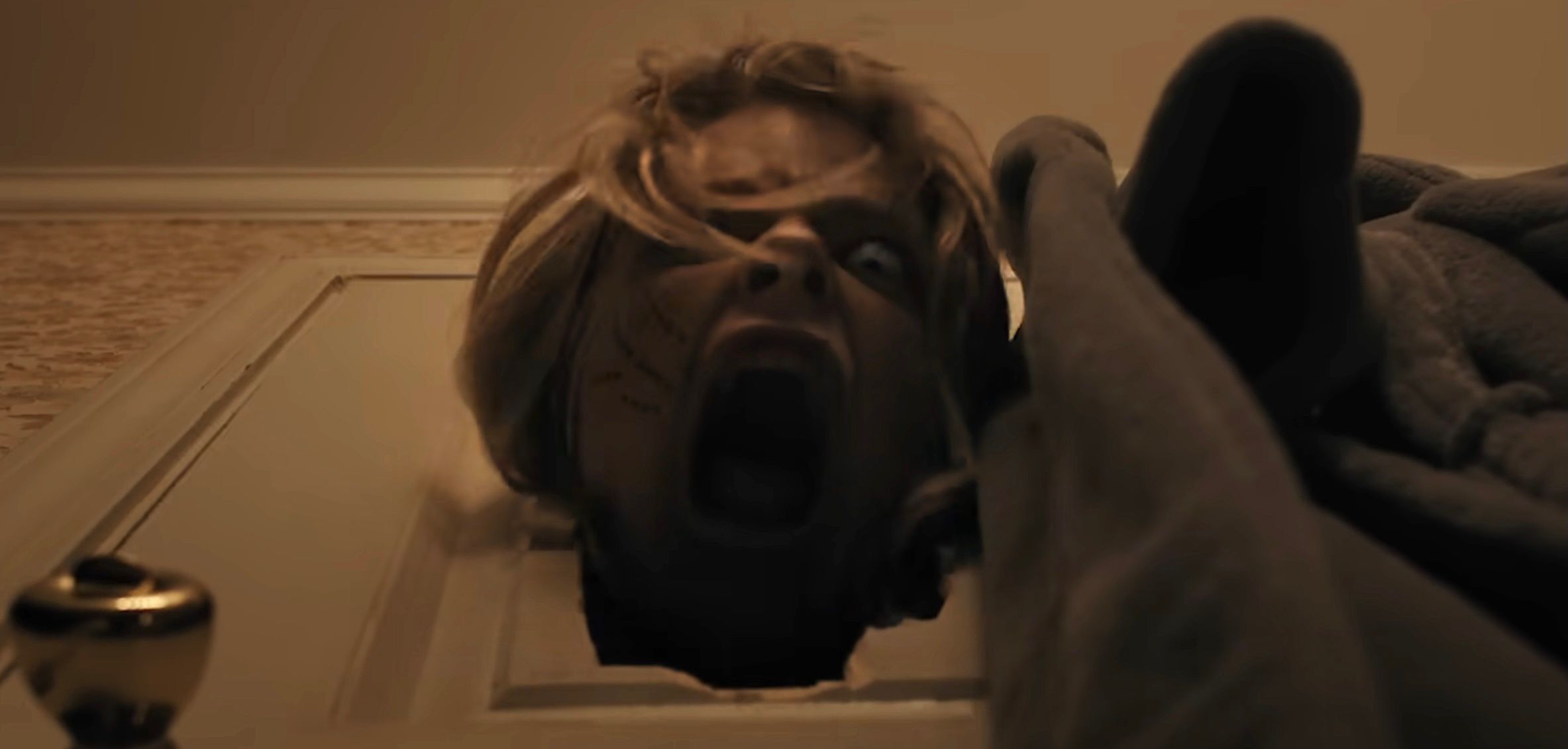 Uma mulher de olhos arregalados grita para baixo em direção à câmera, sua cabeça se casou através de um buraco esmagado em uma porta branca, em armas