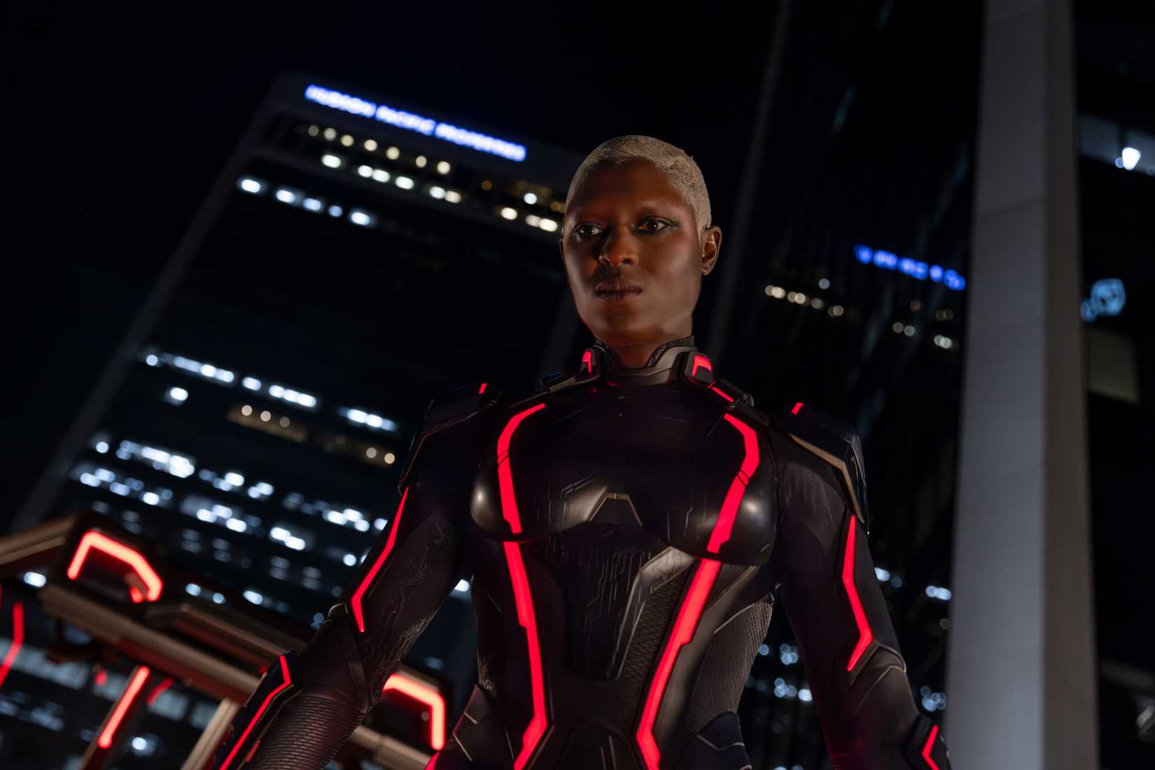 Athena (Jodie Turner-Smith), uma mulher negra com cabelos brancos de malha curta, vestindo uma armadura preta brilhante com destaques brilhantes de neon vermelho, fica em frente a uma janela que se abre para uma paisagem noturna em Tron: Ares