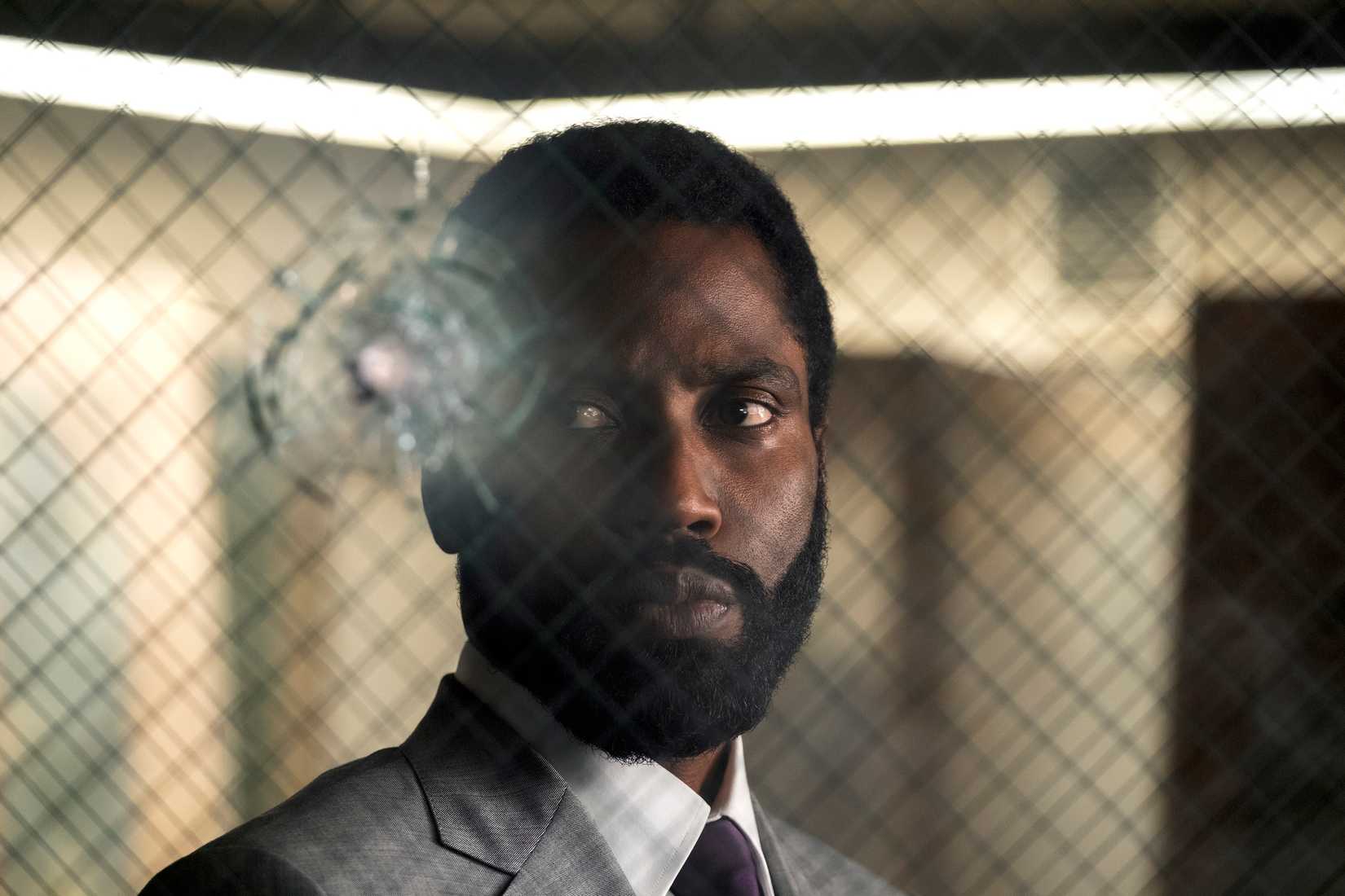 John David Washington em pé em frente a uma ferida de bala em vidro à prova de balas no princípio