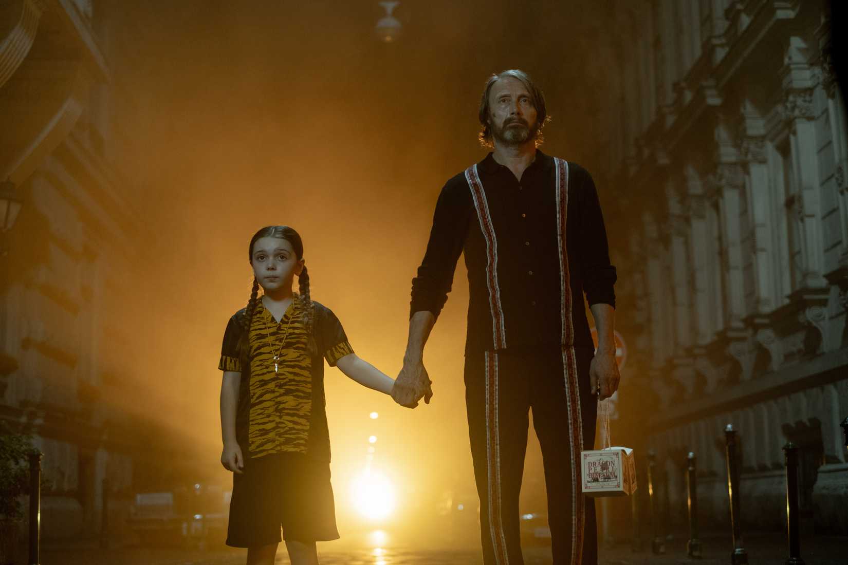 Imagem das atrações na estrada, com Mads Mikkelsen e Sophie Sloan. Os dois dão as mãos enquanto uma luz amarela das luzes de um carro brilha atrás deles. Eles estão em uma rua. 