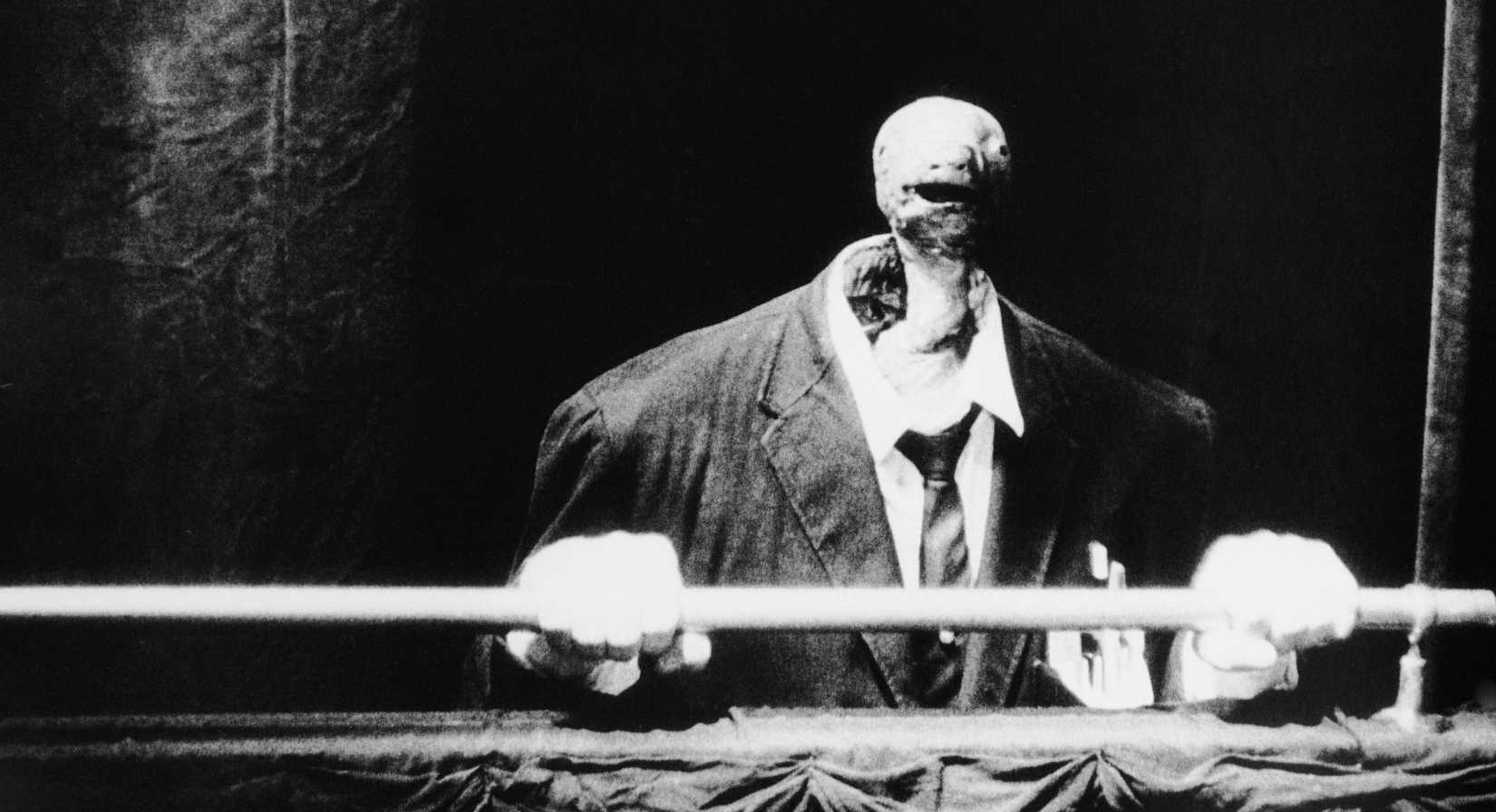Uma figura assustadora que consiste em um terno e braços de homem, com as mãos segurando um corrimão e uma cabeça fina de cobra emergindo do colarinho em vez de uma cabeça humana, em Eraserhead