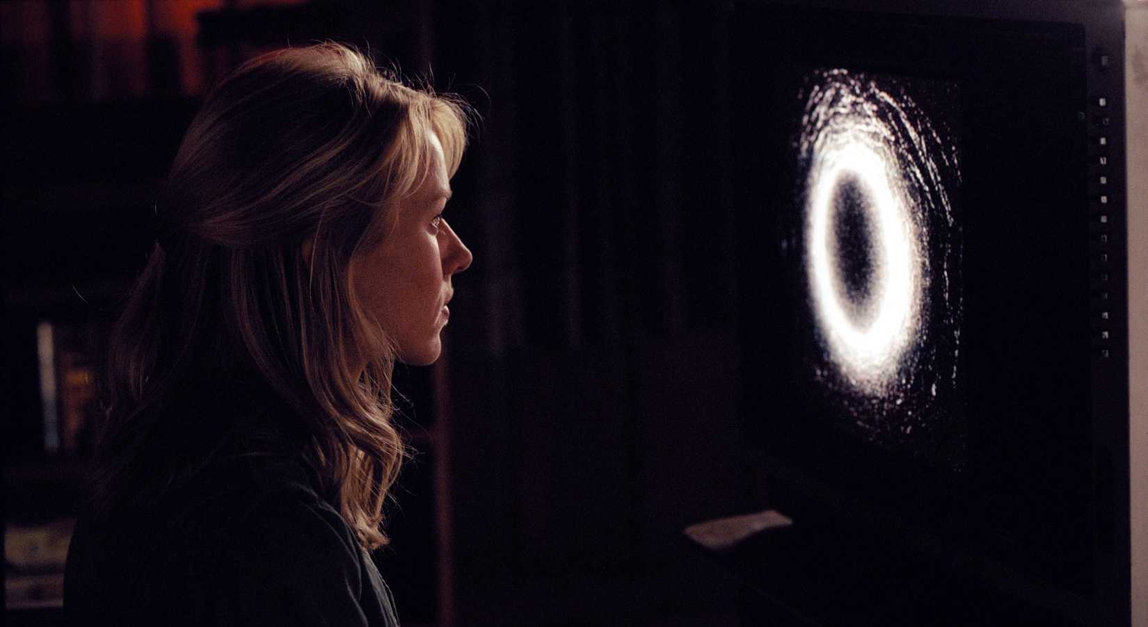 Naomi Watts está sentada em um quarto escuro, olhando para um anel branco brilhante em uma tela de TV em The Ring