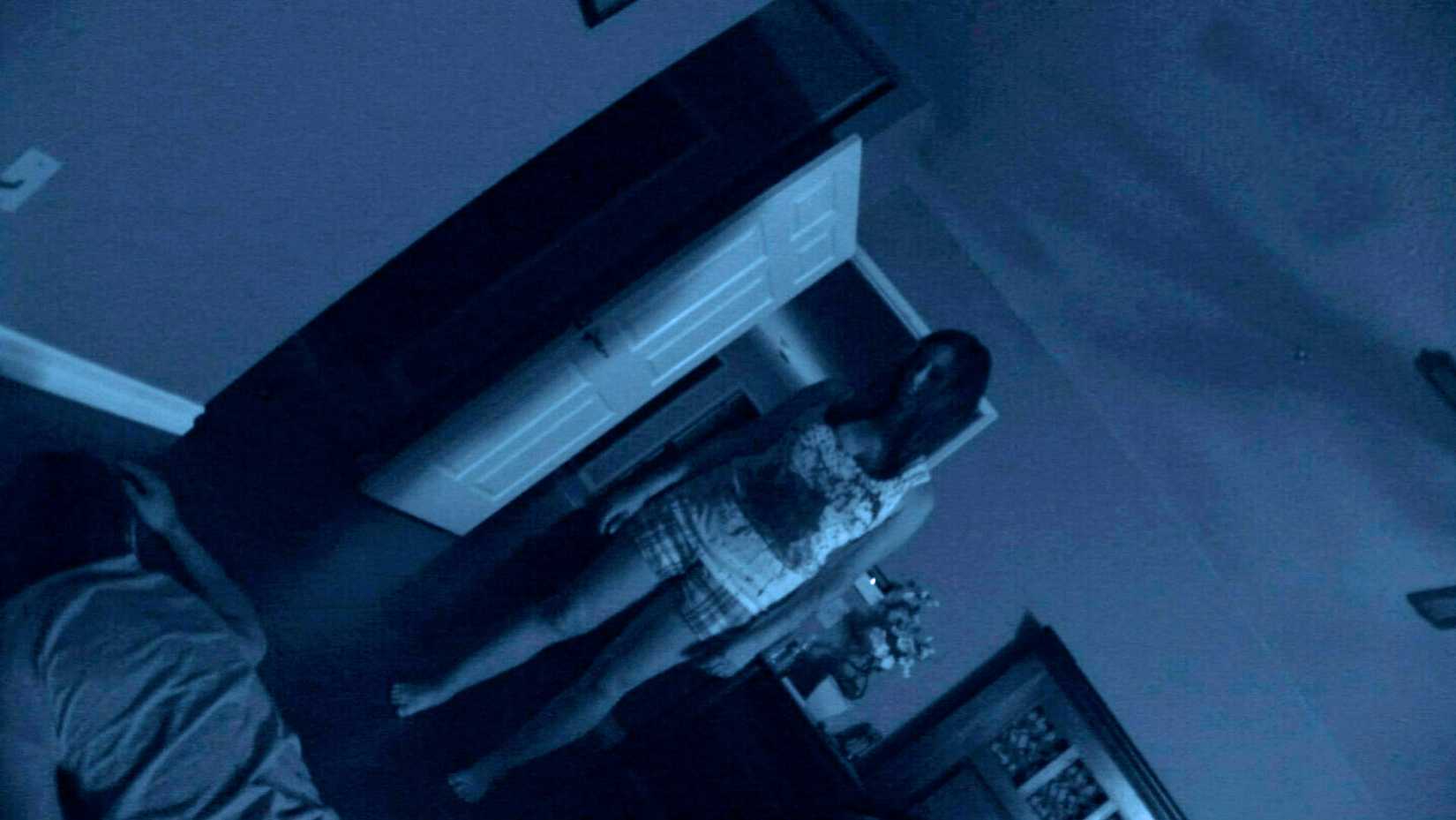 Uma foto monocromática em inclinação holandesa de Atividade Paranormal de 2007, mostrando uma visão mal iluminada de uma mulher de camiseta e shorts xadrez com uma enorme mancha na barriga em pé sobre um corpo de próstata