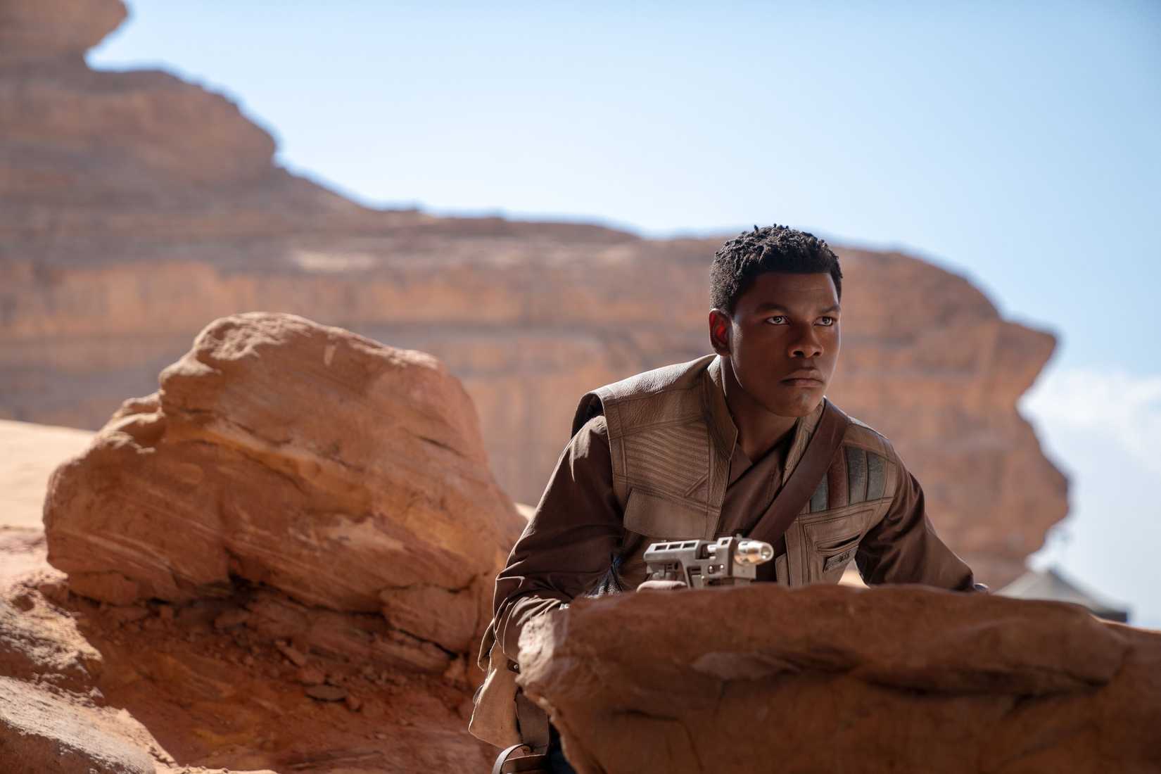 Imagem de John Boyega como Finn em The Rise of Skywalker. Ele está cercado por um local deserto e se esconde entre as rochas.
