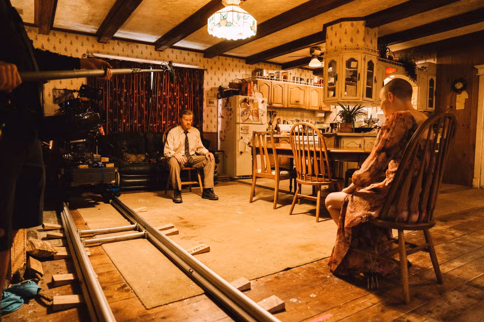 Nos bastidores do set de Bugonia, com Jesse Plemons e Emma Stone sentados em cadeiras de madeira frente a frente enquanto uma câmera sobre trilhos observa