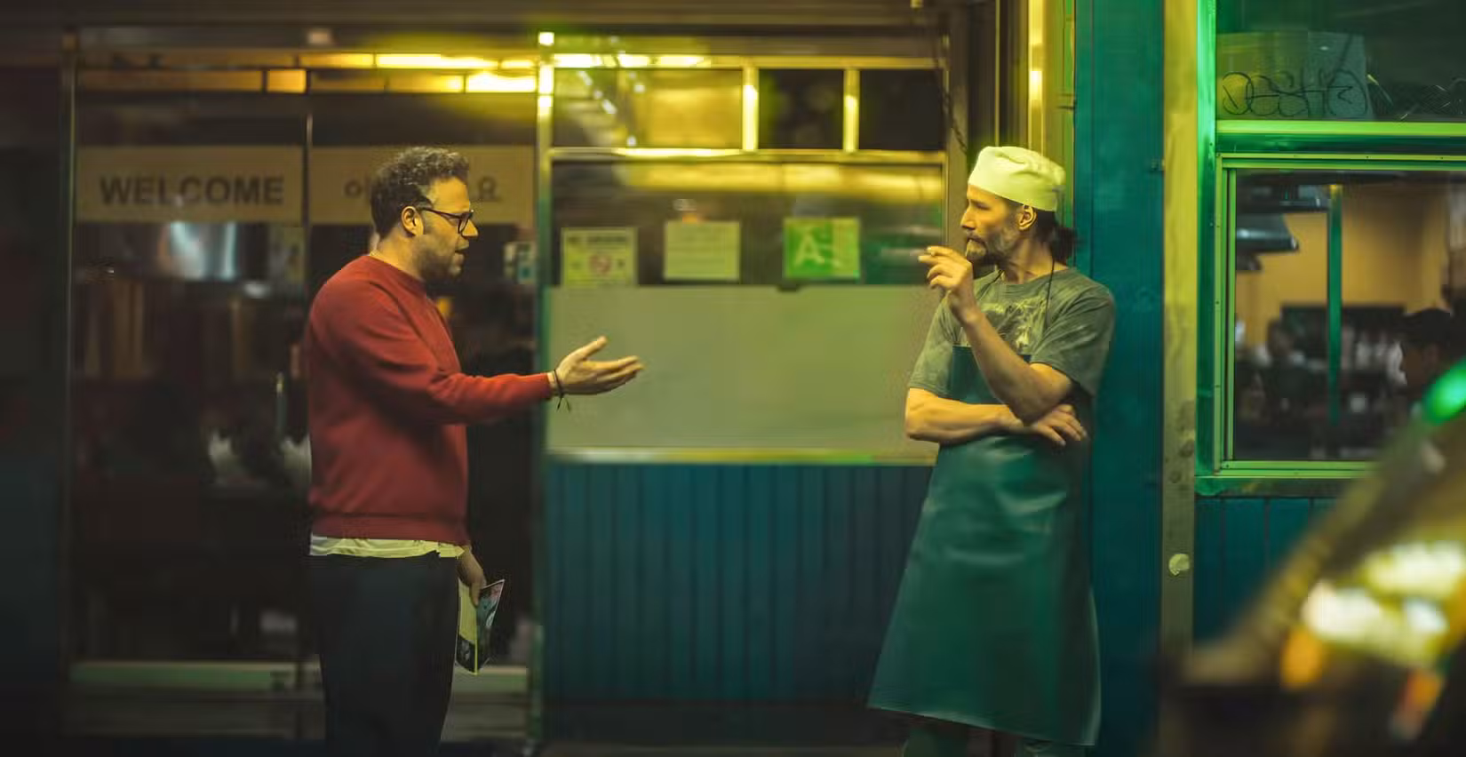 Jeff (Seth Rogen) confronta Gabriel (Keanu Reeves) sob a luz verde nada lisonjeira de um restaurante coreano em um shopping center. Reeves está com o cabelo preso em um rabo de cavalo, usando avental e boné de papel para serviço de alimentação e fumando.