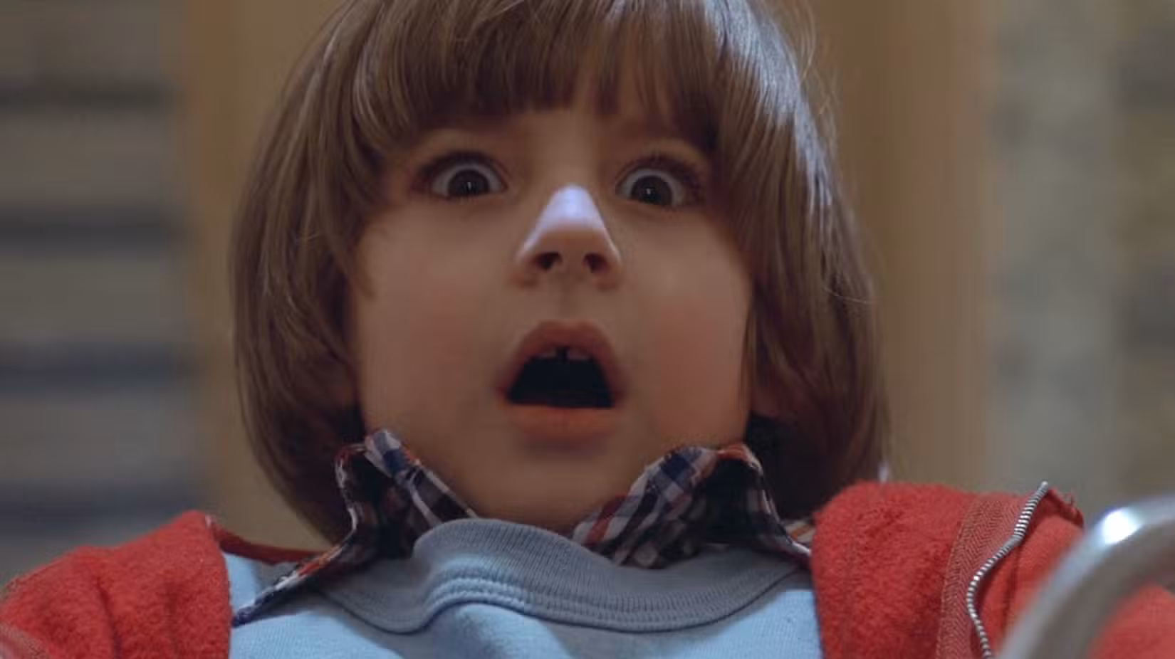 Um close de Danny Torrance (Danny Lloyd), uma criança muito pequena com cabelo cortado em tigela e uma jaqueta vermelha, reagindo com horror de olhos arregalados e boca aberta às gêmeas fantasmas na adaptação cinematográfica de Stanley Kubrick de O Iluminado