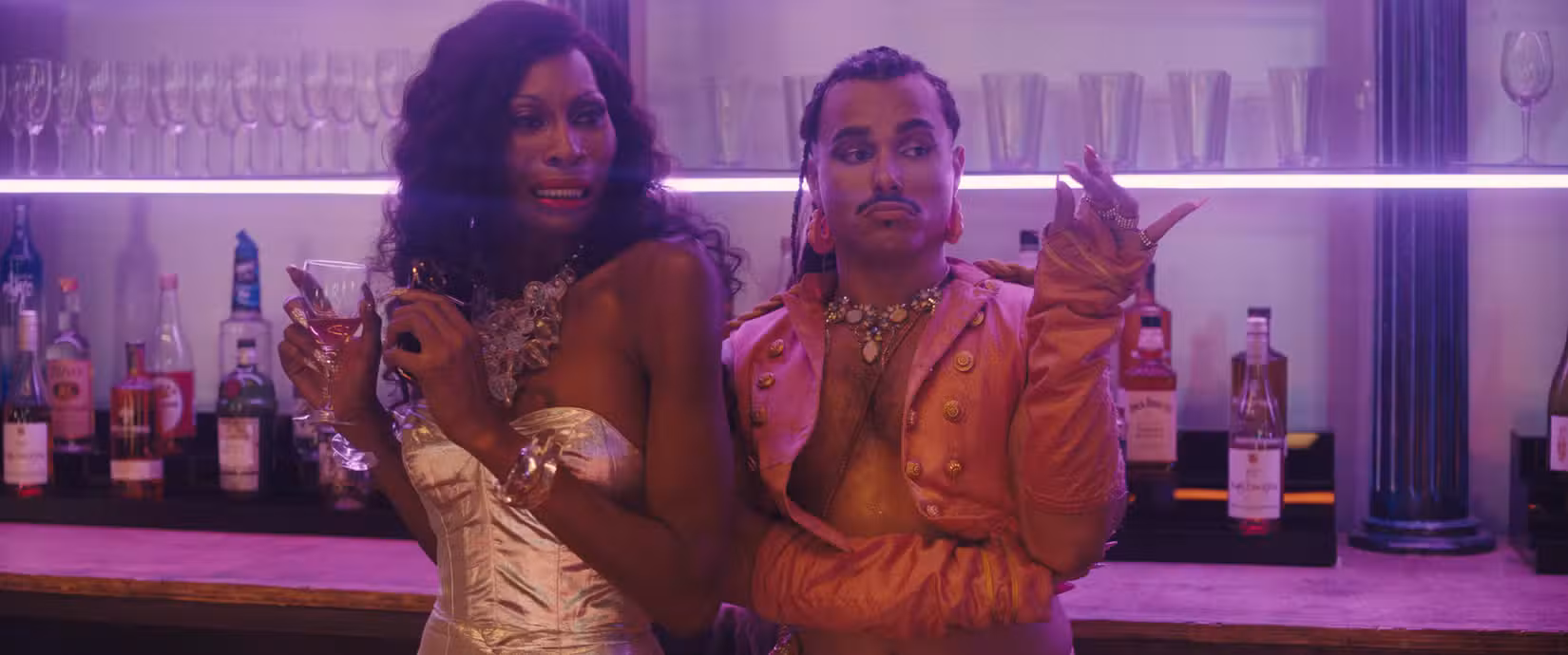 Uma mulher negra com um corpete prateado (Dominique Jackson) e um homem sem camisa com um colete aberto de couro salmão de mangas compridas (Tomás Matos) estão em um bar, olhando de soslaio para algo fora da tela, em Queens of the Dead