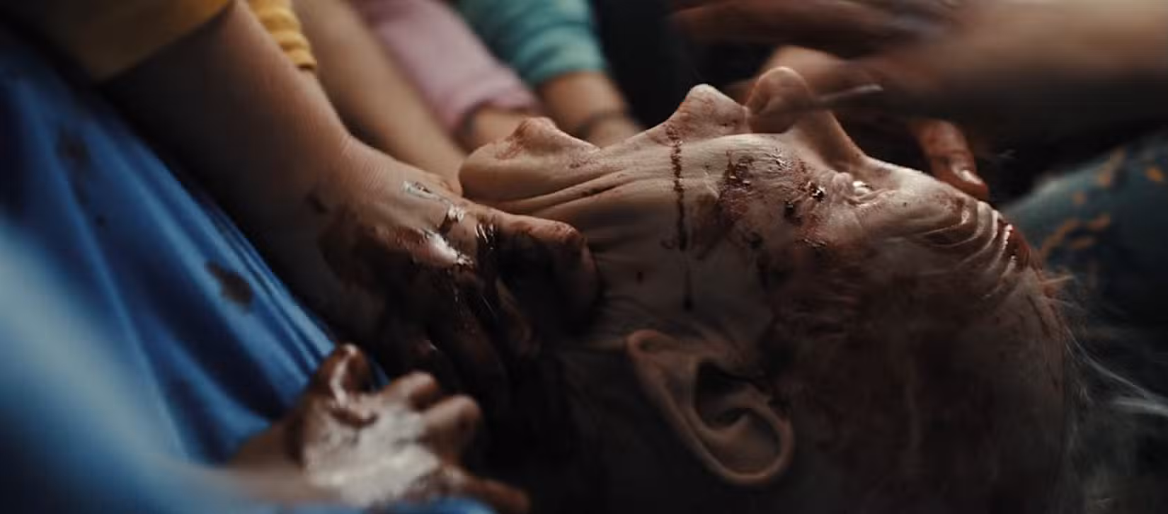 Um close do rosto ensanguentado e gritando de uma mulher enquanto uma multidão de crianças a ataca por todos os lados em Armas