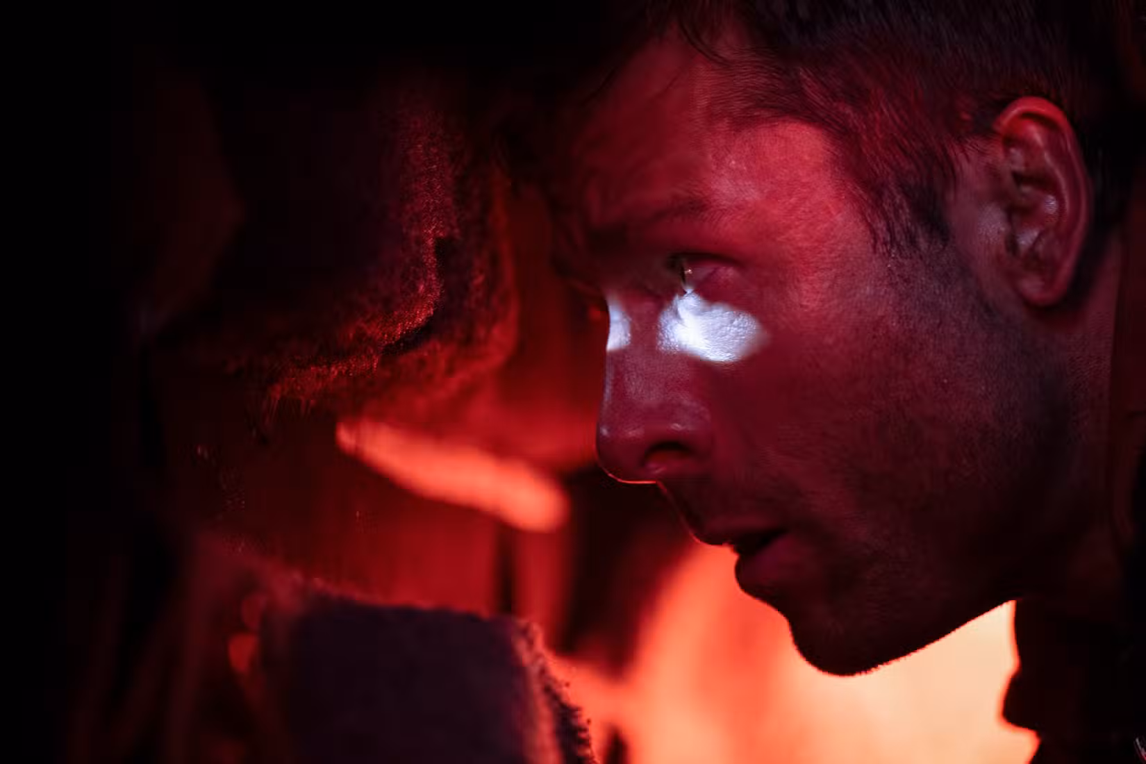 Ben Richards (Glen Powell), em um porta-malas escuro e iluminado em vermelho, espia através de um buraco de bala para o exterior na adaptação de 2025 de The Running Man, de Stephen King