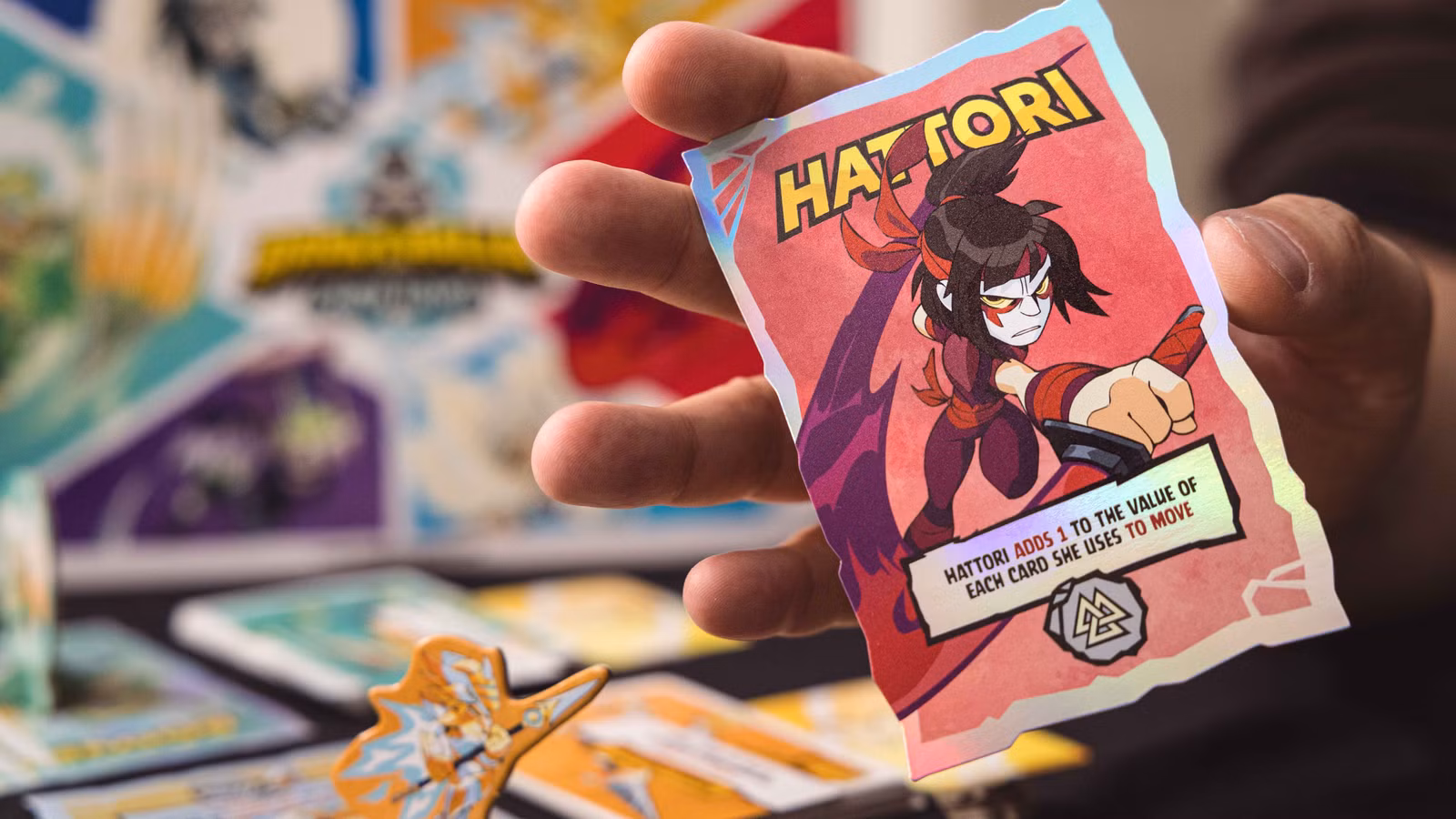 Uma carta representando Hattori mostrada na frente de uma imagem com a caixa e componentes de Brawlhalla: The Card Game