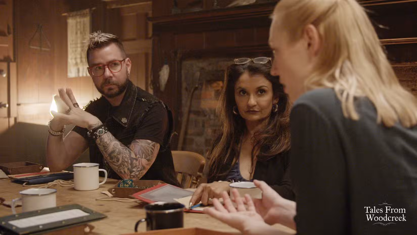 Kelly McLaughlin e Anjali Bhimani sentam-se em uma mesa olhando para a Dungeon Master Deborah Ann Woll durante um episódio de Tales From Woodcreek