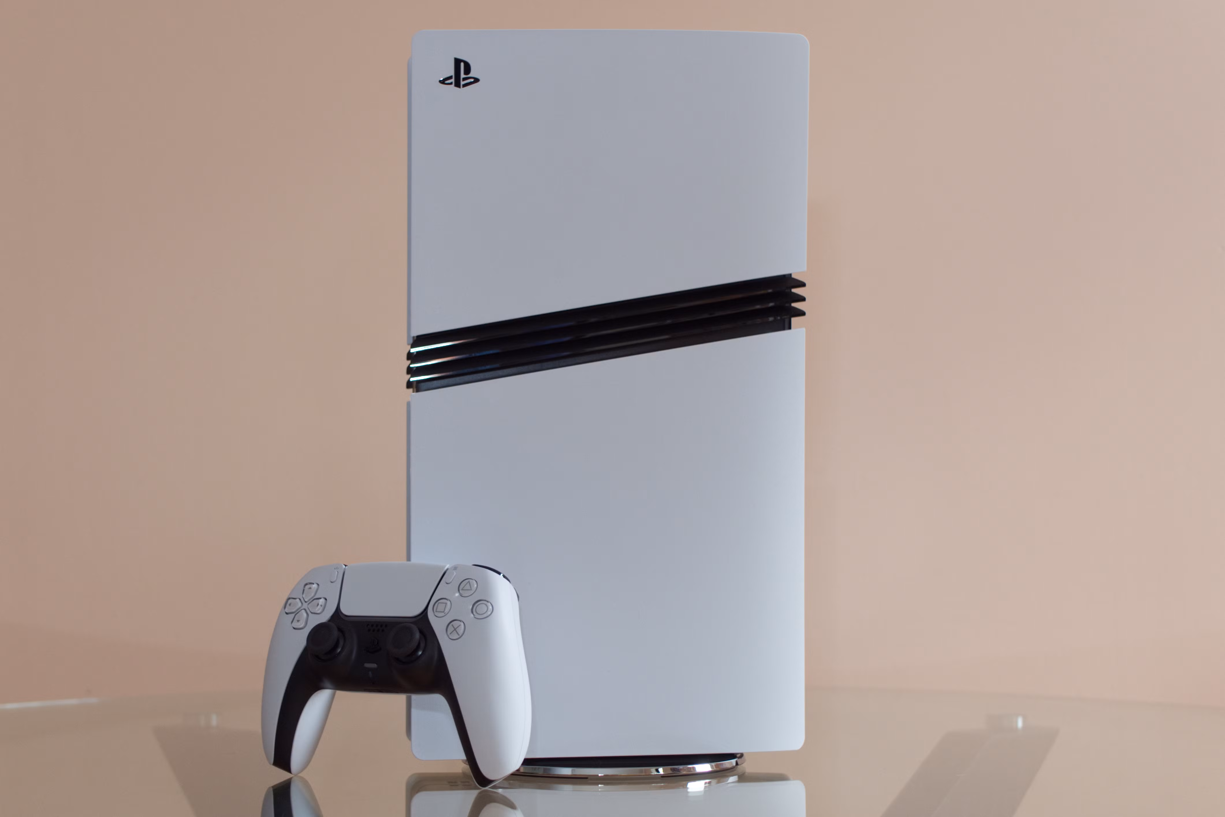 Uma foto do console PlayStation 5 Pro apoiado em uma mesa de vidro, encostada em uma parede cor de pêssego. O controlador DualSense está encostado no console.