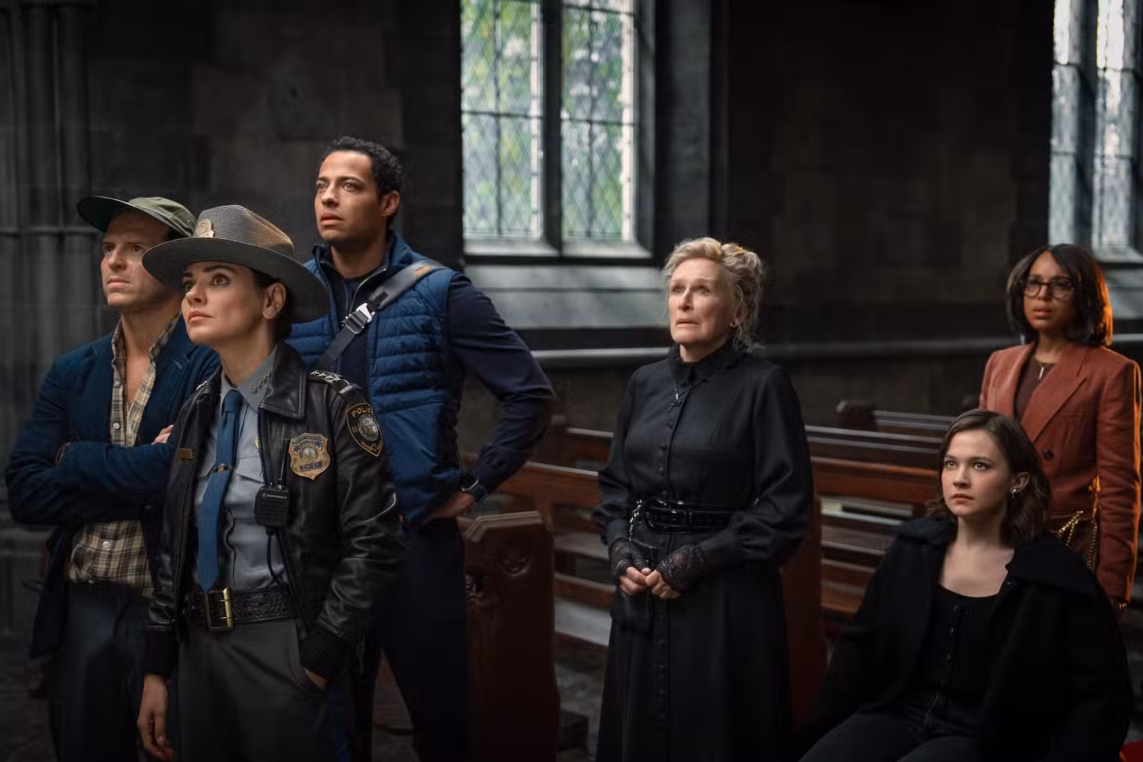 Em uma cena de Wake Up Dead Man, fiéis da igreja interpretados por Glenn Close, Daryl McCormack, Andrew Scott, Cailee Spaeny e Kerry Washington, bem como uma policial interpretada por Mila Kunis, reúnem-se nos bancos de uma igreja.