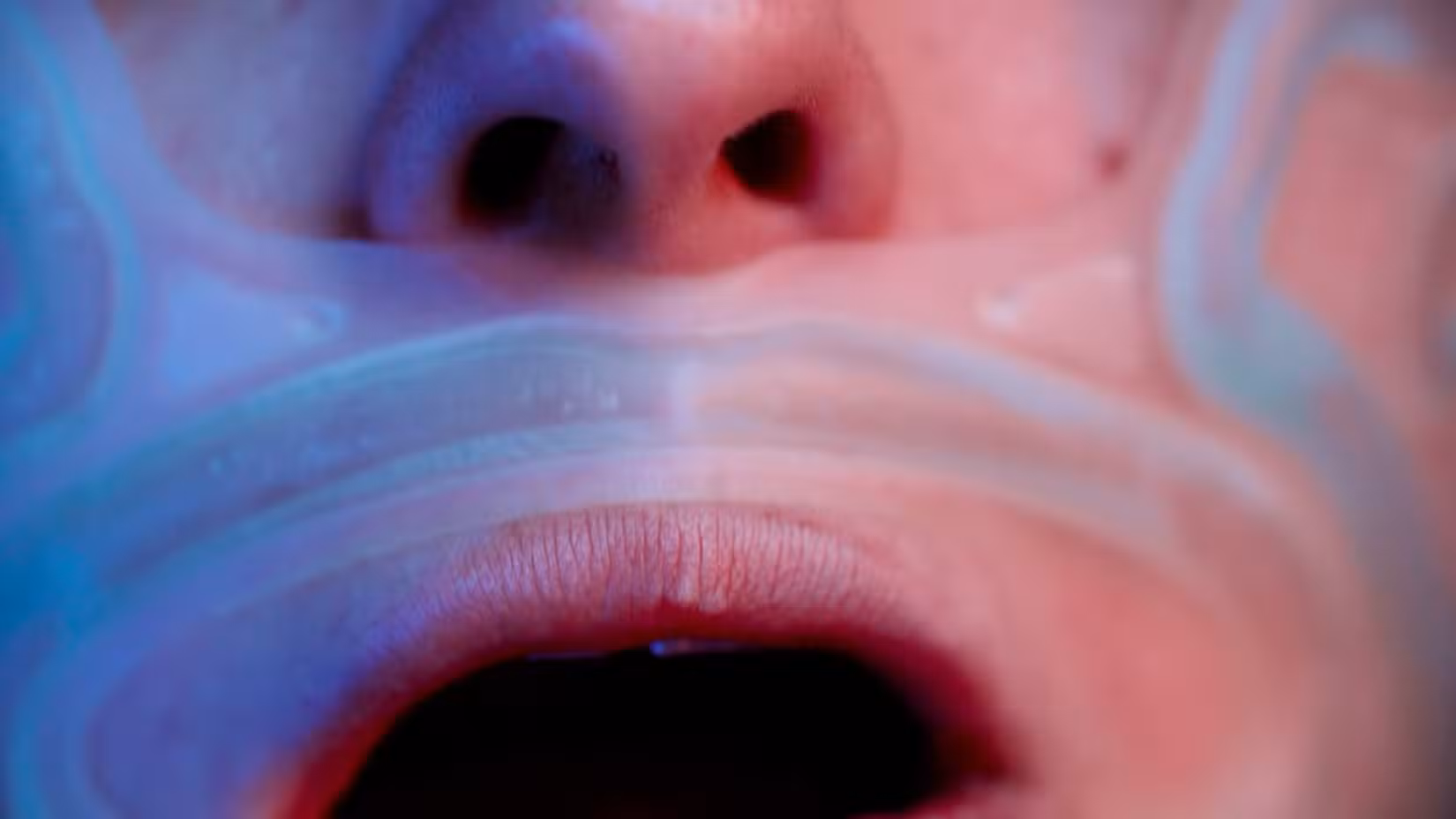 Um close de uma boca humana, aberta, com algum tipo de substância translúcida e iridescente espalhada sobre partes da pele da pessoa.