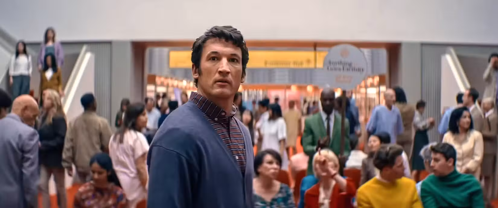 Larry (Miles Teller) fica olhando para a câmera, com uma enorme multidão de pessoas fora de foco atrás dele, na Eternidade