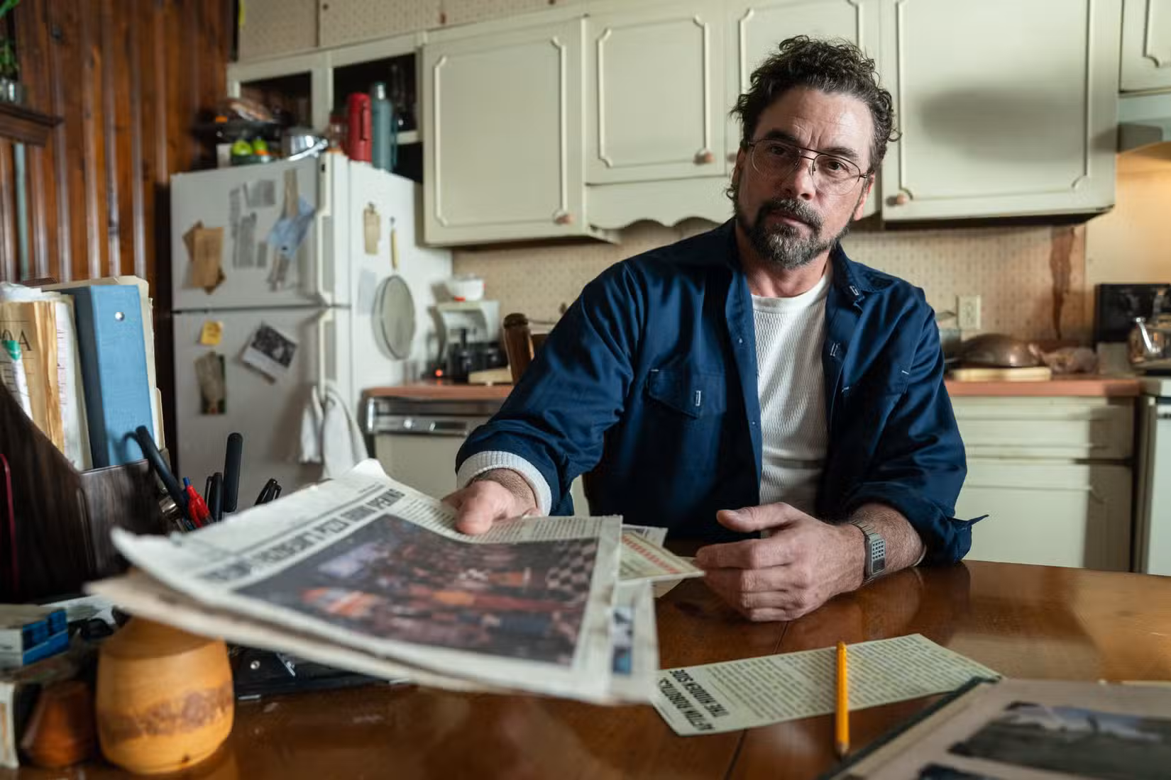 Skeet Ulrich está sentado à mesa de uma pequena cozinha, segurando um jornal em direção à câmera, em Five Nights at Freddy's 2