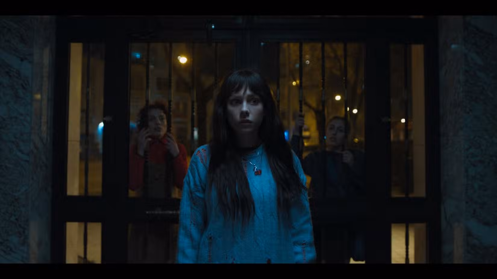 Uma jovem de cabelos castanhos está em frente à entrada de um prédio de apartamentos. Ela usa um suéter azul e um colar. Está escuro e ela parece nervosa.