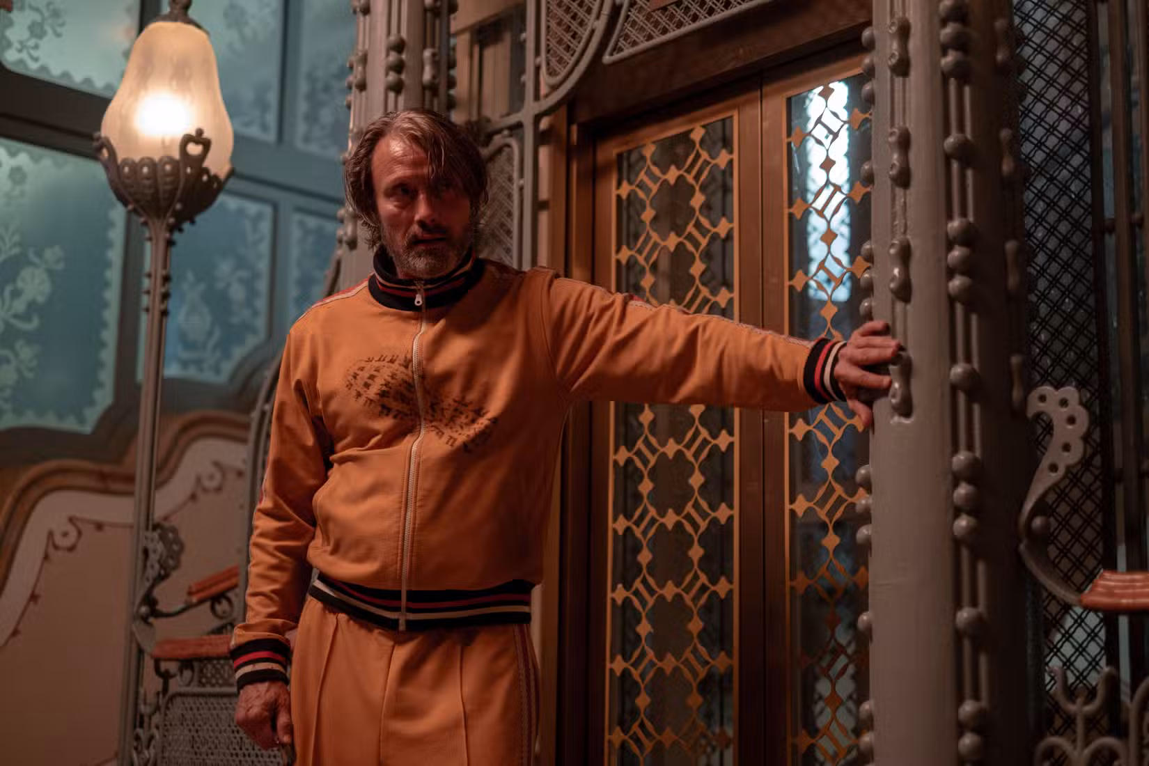 Uma imagem do Dust Bunny de Bryan Fuller. Mads Mikkelsen veste um agasalho amarelo e se encosta em um elevador.