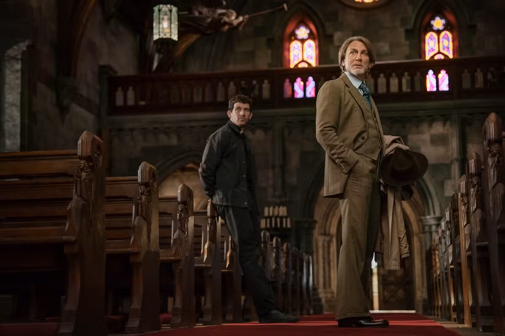 Dois homens (Josh O'Connor e Daniel Craig) estão no corredor de uma igreja católica, com vitrais acima deles, em Wake Up Dead Man