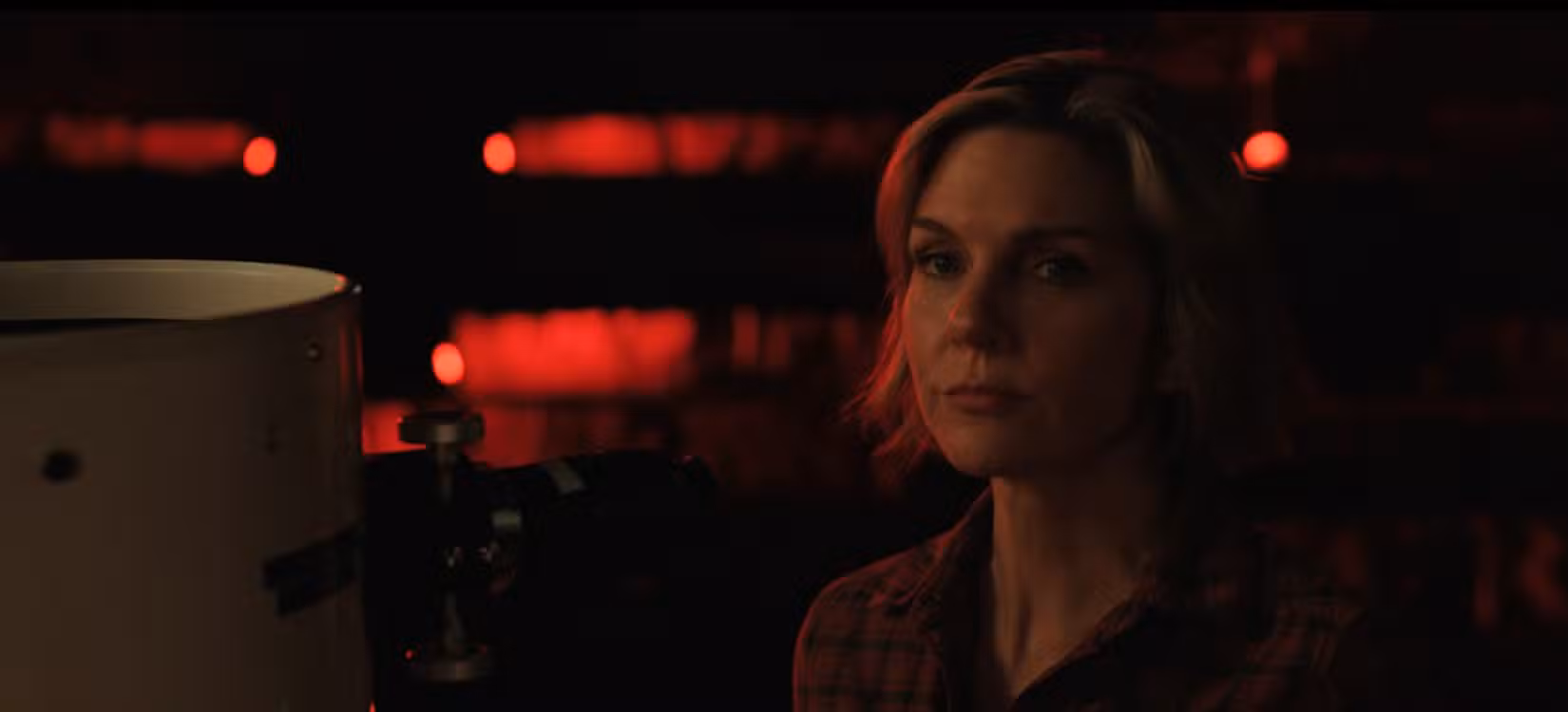 Carol (Rhea Seehorn) parece irônica em um espaço escuro e vermelho, parada ao lado de um telescópio, em Pluribus