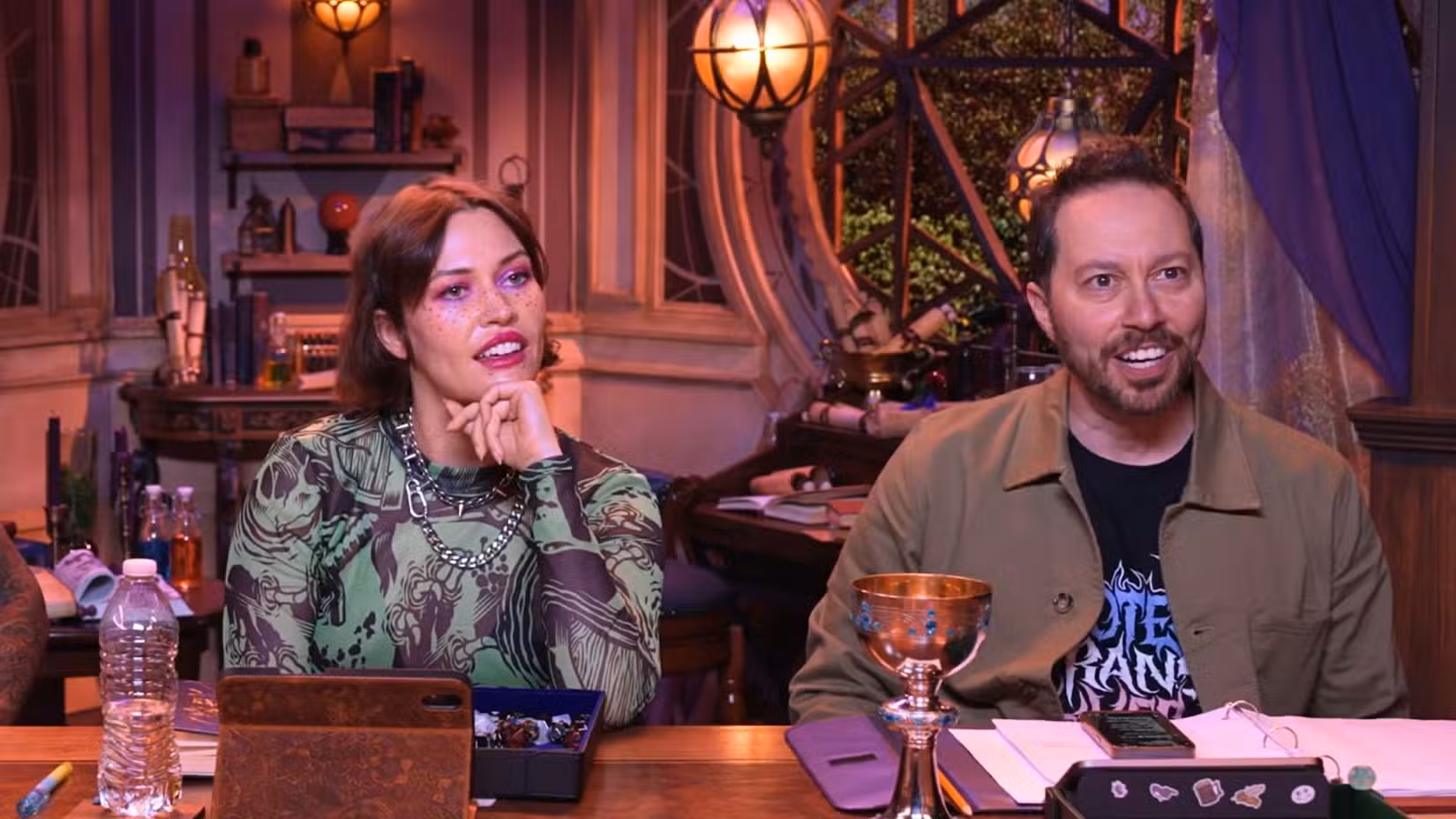 Uma imagem do Cooldown do Critical Role. Mostra Whitney Moore e Sam Riegel sentados um ao lado do outro em uma mesa de D&D.