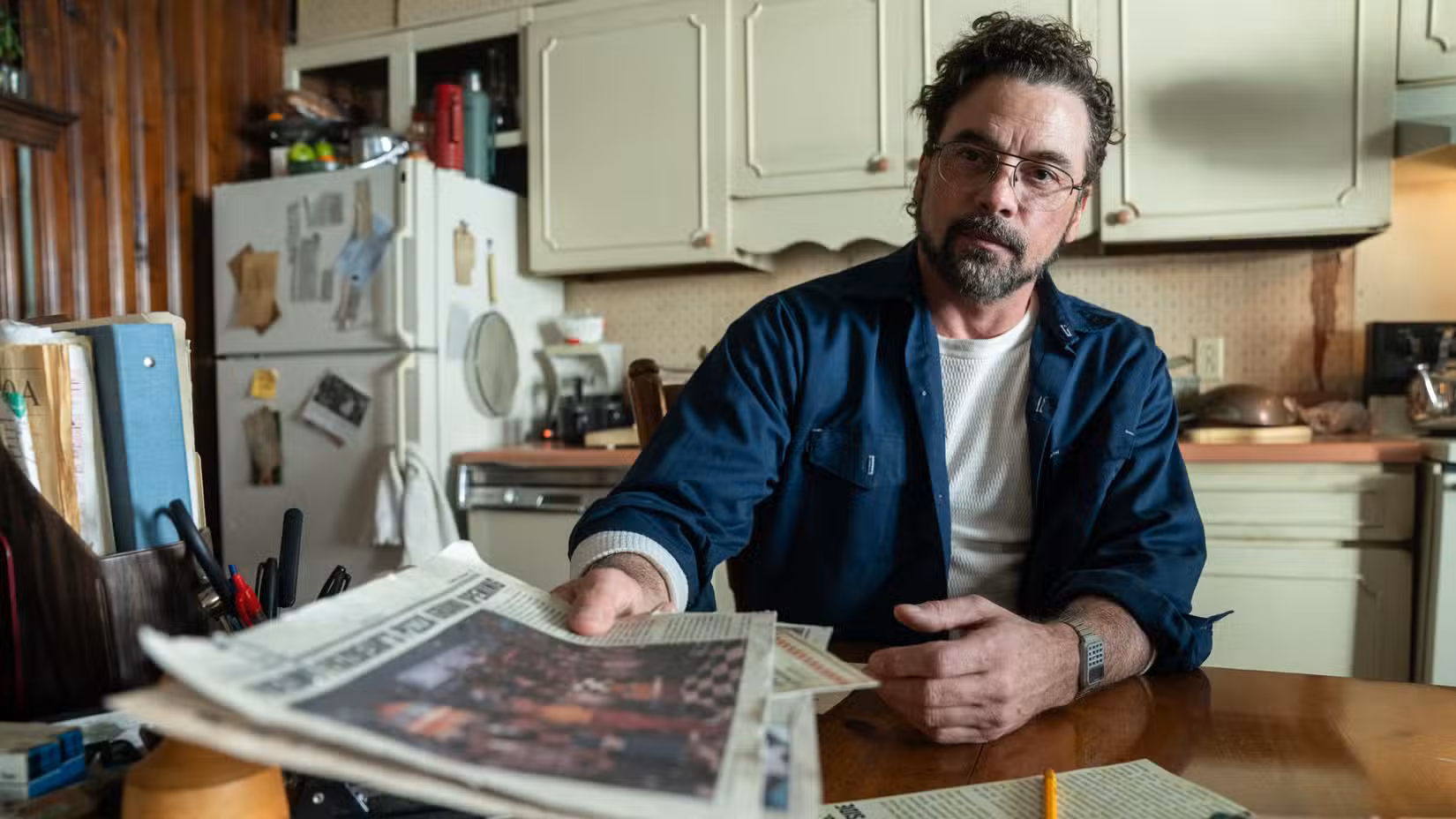 Skeet Ulrich está sentado à mesa de uma pequena cozinha, segurando um jornal em direção à câmera, em Five Nights at Freddy's 2