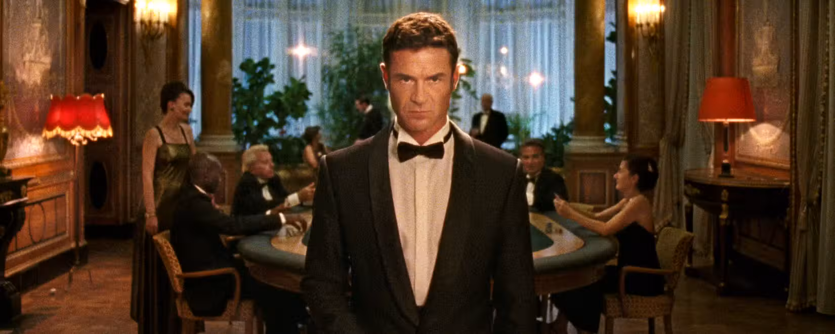 Um homem de aparência severa e smoking (Yannick Renier) está parado no meio da tela, olhando atentamente para a câmera. Atrás dele, homens de terno e mulheres em vestidos de festa jogam cartas em uma mesa de cassino.