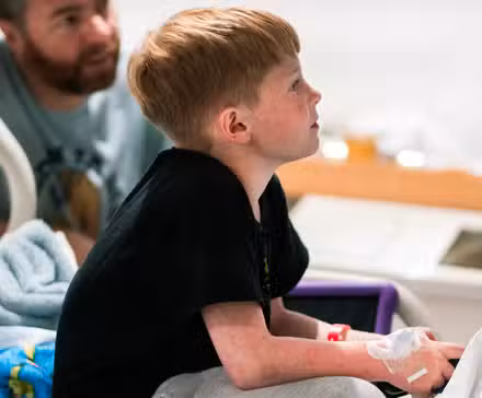Um garotinho loiro com tubos presos ao braço joga videogame em uma cama de hospital