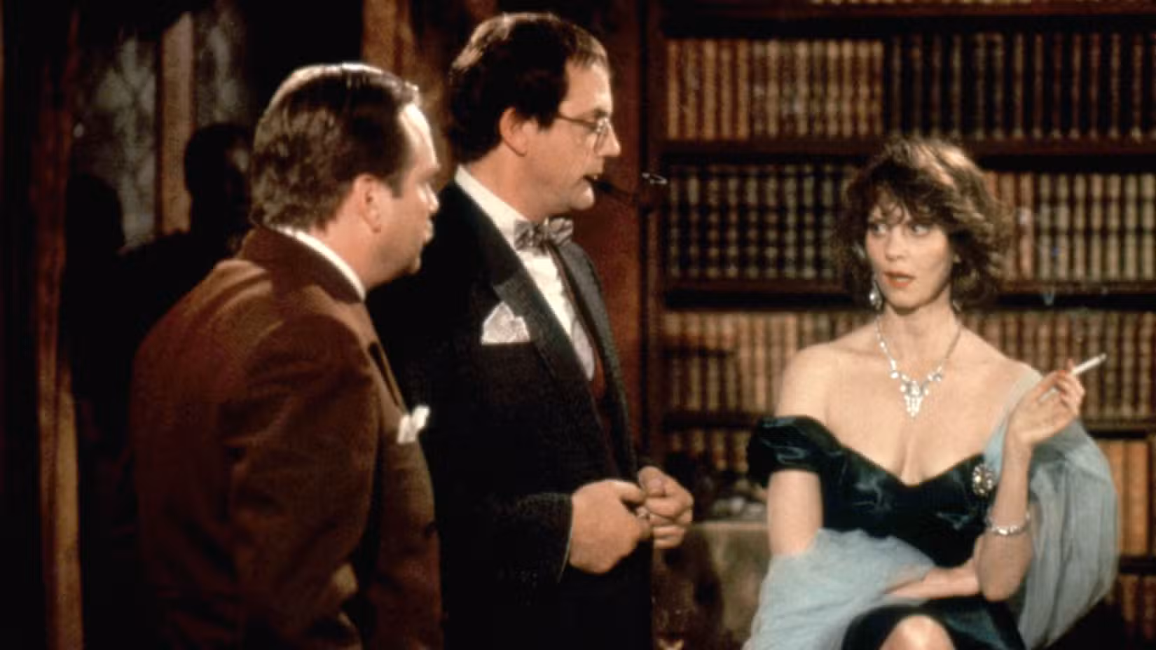 Miss Scarlet (Lesley Ann Warren) fuma um cigarro enquanto é encarada pelo Professor Plum (Christopher Lloyd) e pelo Coronel Mustard (Martin Mull)