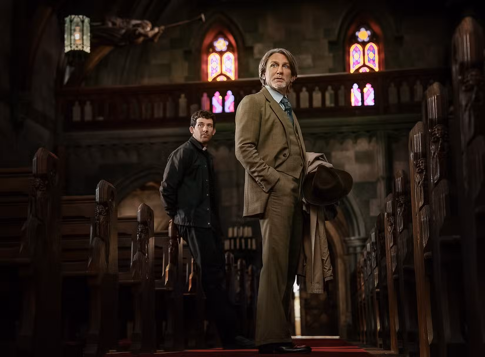 Daniel Craig, como o detetive Benoit Blanc, e Josh O'Connor, como um padre suspeito de assassinato, olham para o corredor de uma igreja católica em uma cena de Wake Up Dead Man: A Knives Out Mystery.