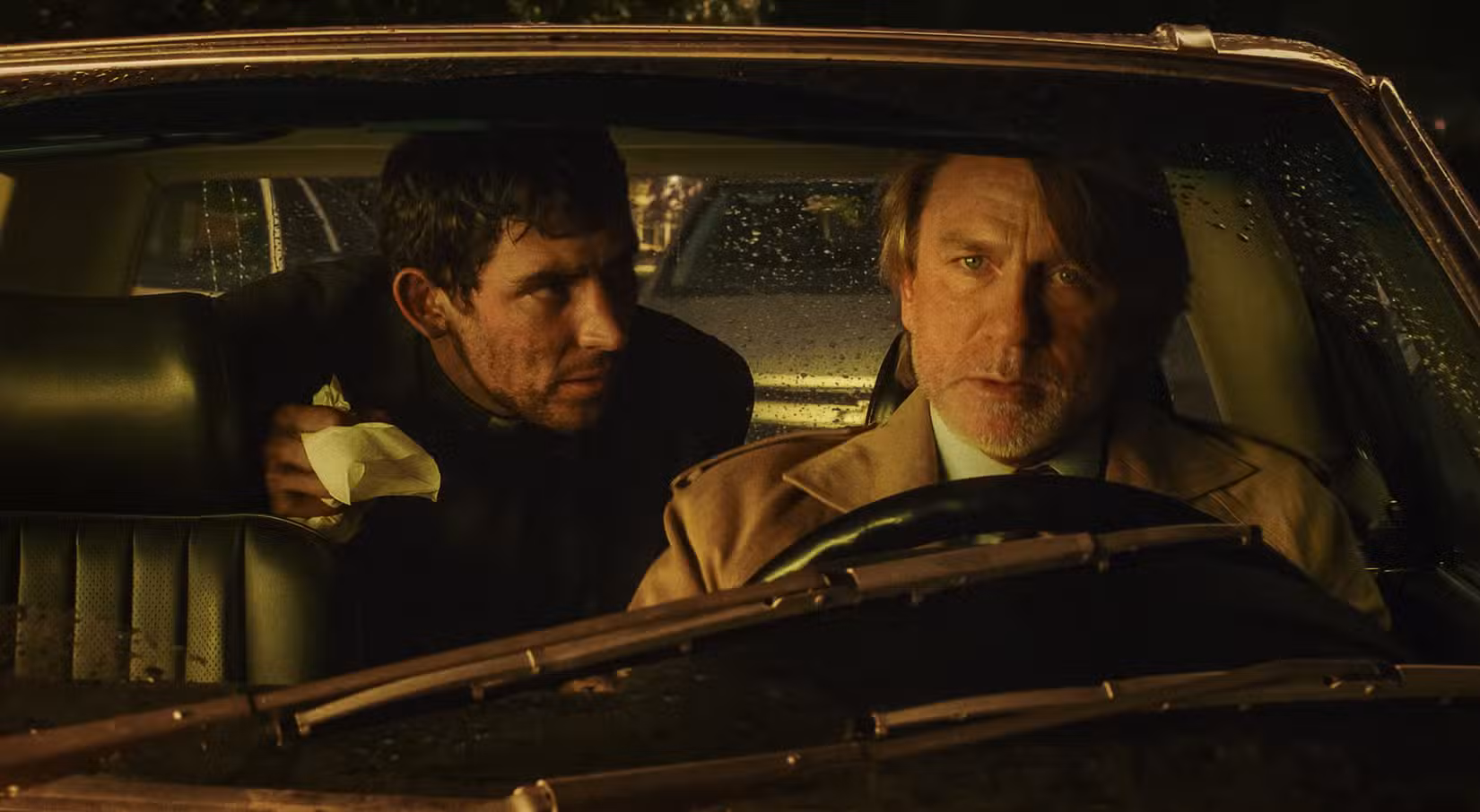 Um padre coberto de sangue (Josh O'Connor) se inclina para frente no banco de trás de um carro à noite para falar com Benoit Blanc (Daniel Craig), que está no banco do motorista, em Wake Up Dead Man