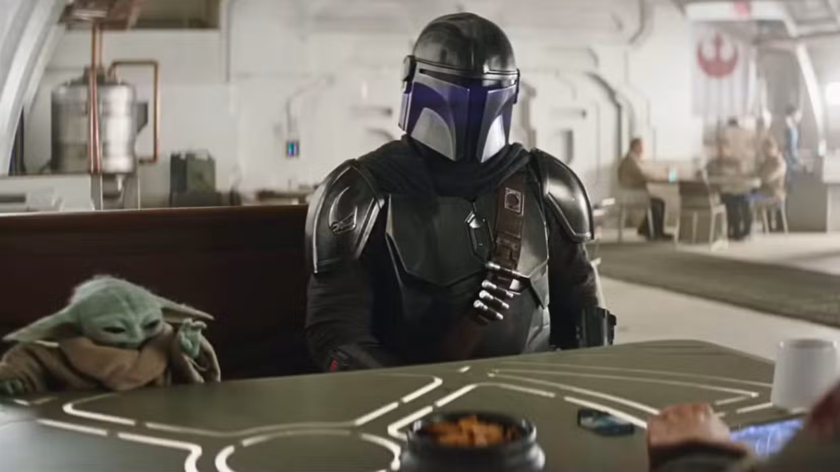 Uma imagem de The Mandalorian e Grogu. Grogu e Din Djarin estão sentados à mesa, com a mão de Grogu estendida.