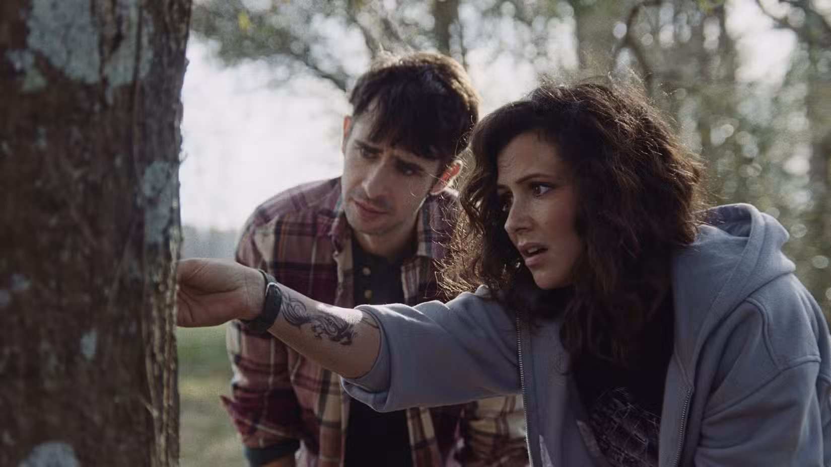 Uma imagem de The Confession, de Will Canon. Italia Ricci e Scott Mechlowicz estão do lado de fora. Ambos estão inclinados para a frente para olhar o tronco de uma grande árvore.