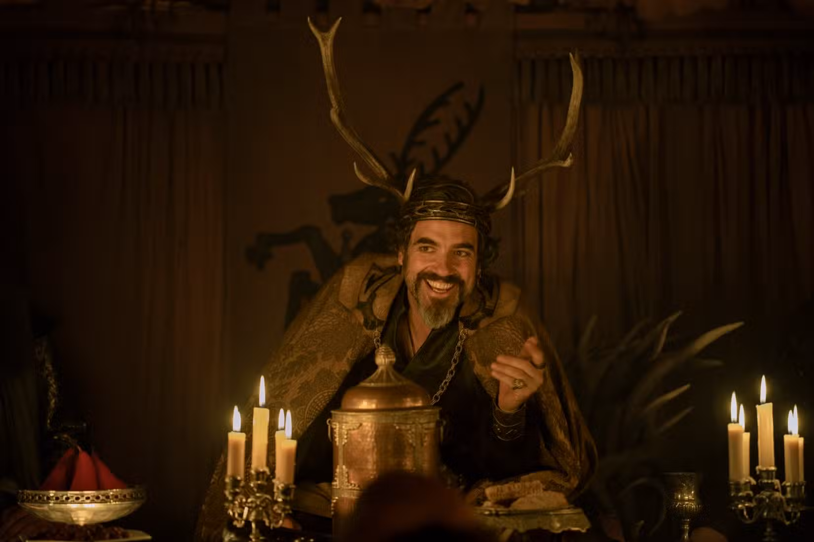Sor Lionel Baratheon (Daniel Ings), com uma capa de couro peluda e um cocar com chifres de veado espalhados, senta-se em uma mesa coberta de velas e sorri em O Cavaleiro dos Sete Reinos