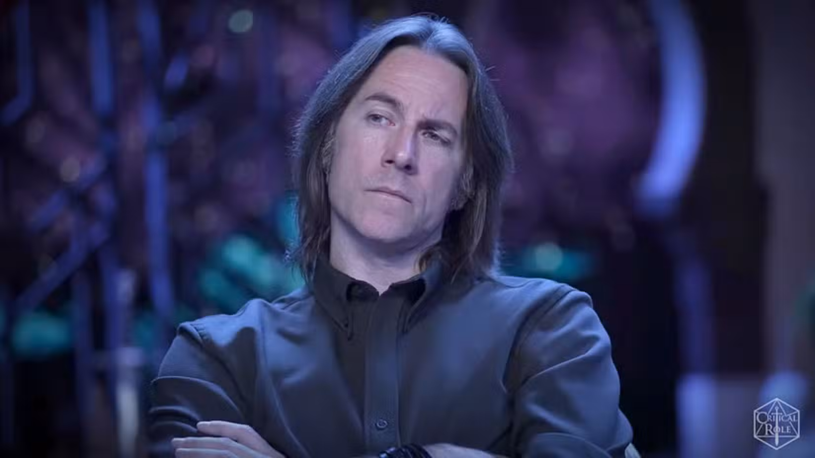 Uma imagem da Critical Role Campaign 4, episódio 12. Apresenta Matthew Mercer, com os braços cruzados sobre o peito, vestindo uma camisa preta.