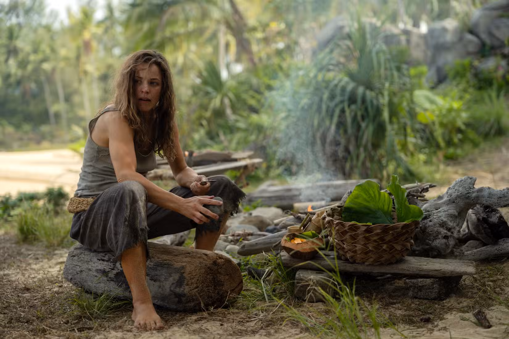 Linda (Rachel McAdams), uma mulher com roupas sujas e sujas, senta-se em um tronco perto de uma fogueira feita à mão entre frutas colhidas e uma cesta tecida à mão em Send Help, de Sam Raimi.