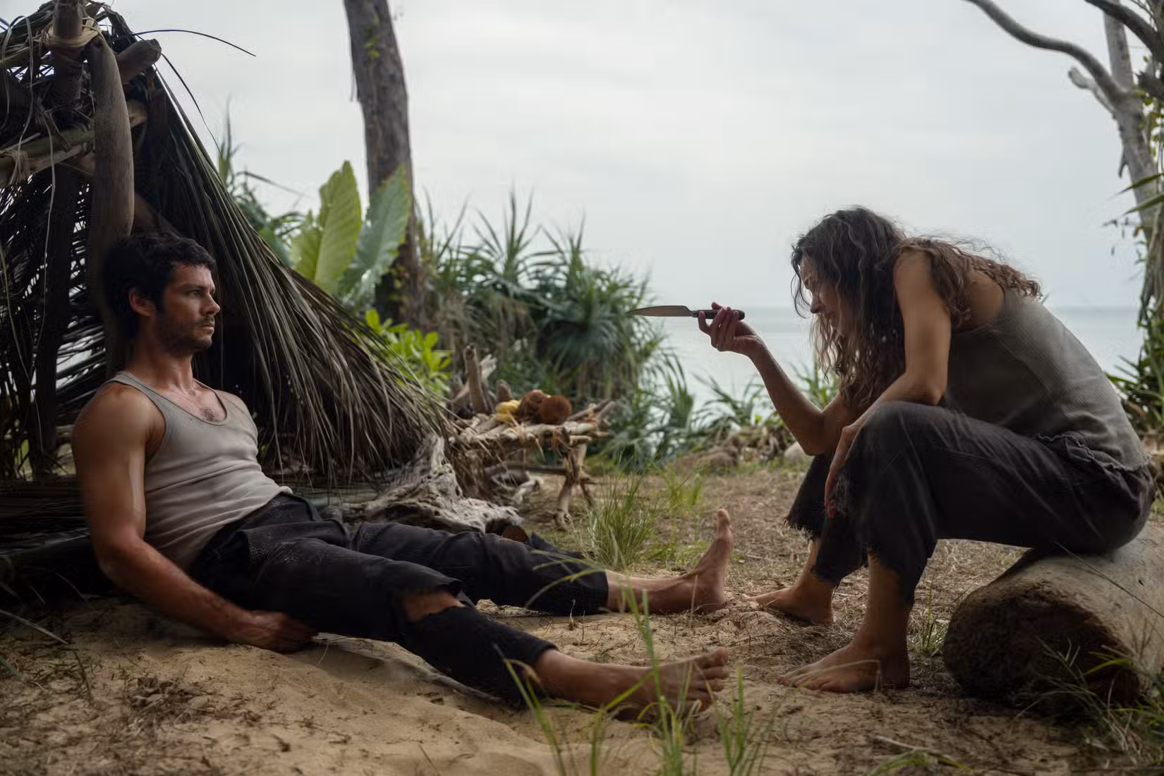 Brad (Dylan O'Brien), um homem descalço e enlameado, com calças rasgadas e uma camiseta imunda sem mangas, se esparrama na areia em frente a um abrigo feito de folhas e folhas, enquanto Linda (Rachel McAdams), uma mulher igualmente suja, gesticula para ele com uma faca em Send Help, de Sam Raimi.