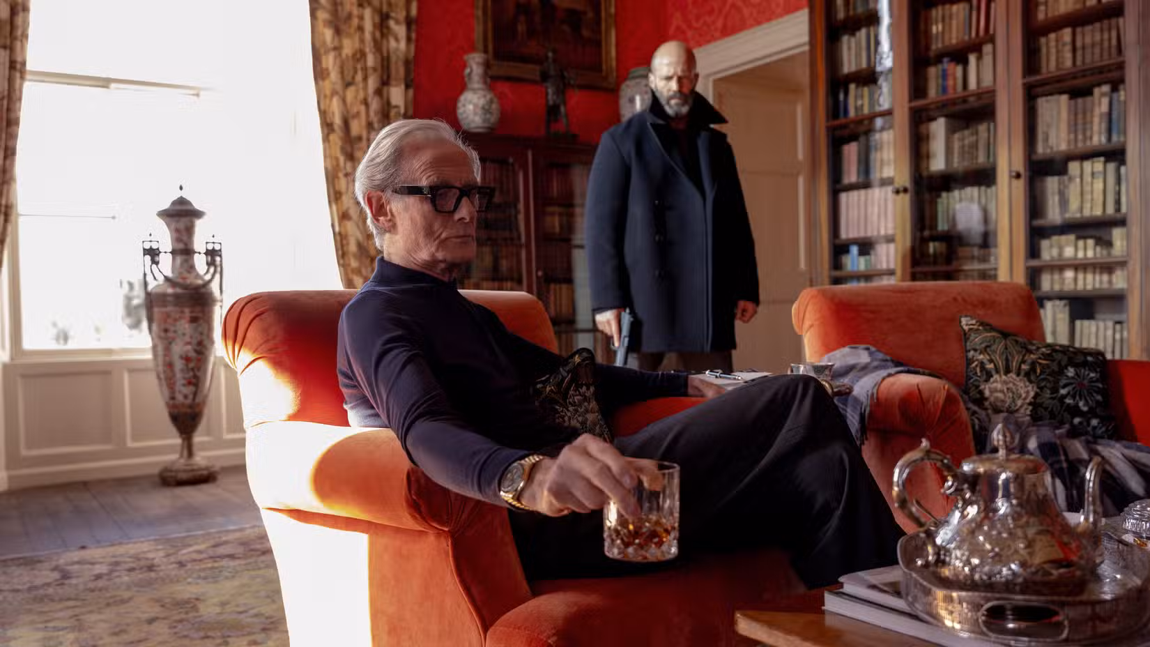 Bill Nighy, como o Big Bad do Shelter, está sentado em uma cadeira de pelúcia laranja brilhante, segurando um coquetel, em uma sala semelhante a uma biblioteca cheia de estantes de livros e vasos elegantes, enquanto Jason Statham aparece atrás dele com uma arma.