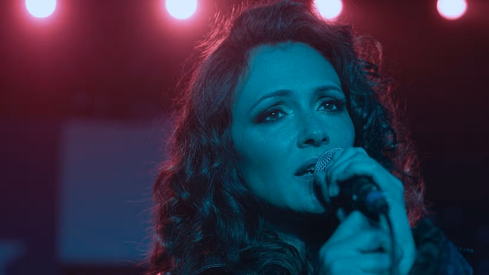 Uma imagem de The Confession, de Will Canon. Italia Ricci enquanto Naomi canta no microfone. A iluminação em seu rosto a faz parecer azul.