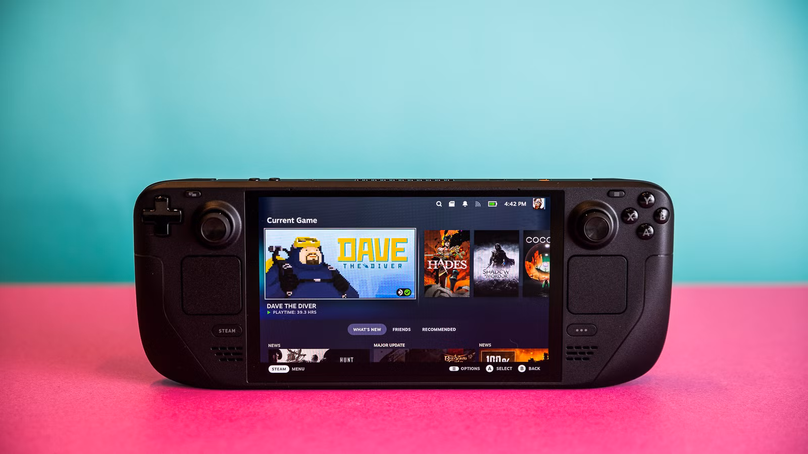 Fotografias do Steam Deck OLED e de seu case rígido com zíper. Cada foto é tirada com o computador de mão sobre uma folha de papel grosso rosa choque.