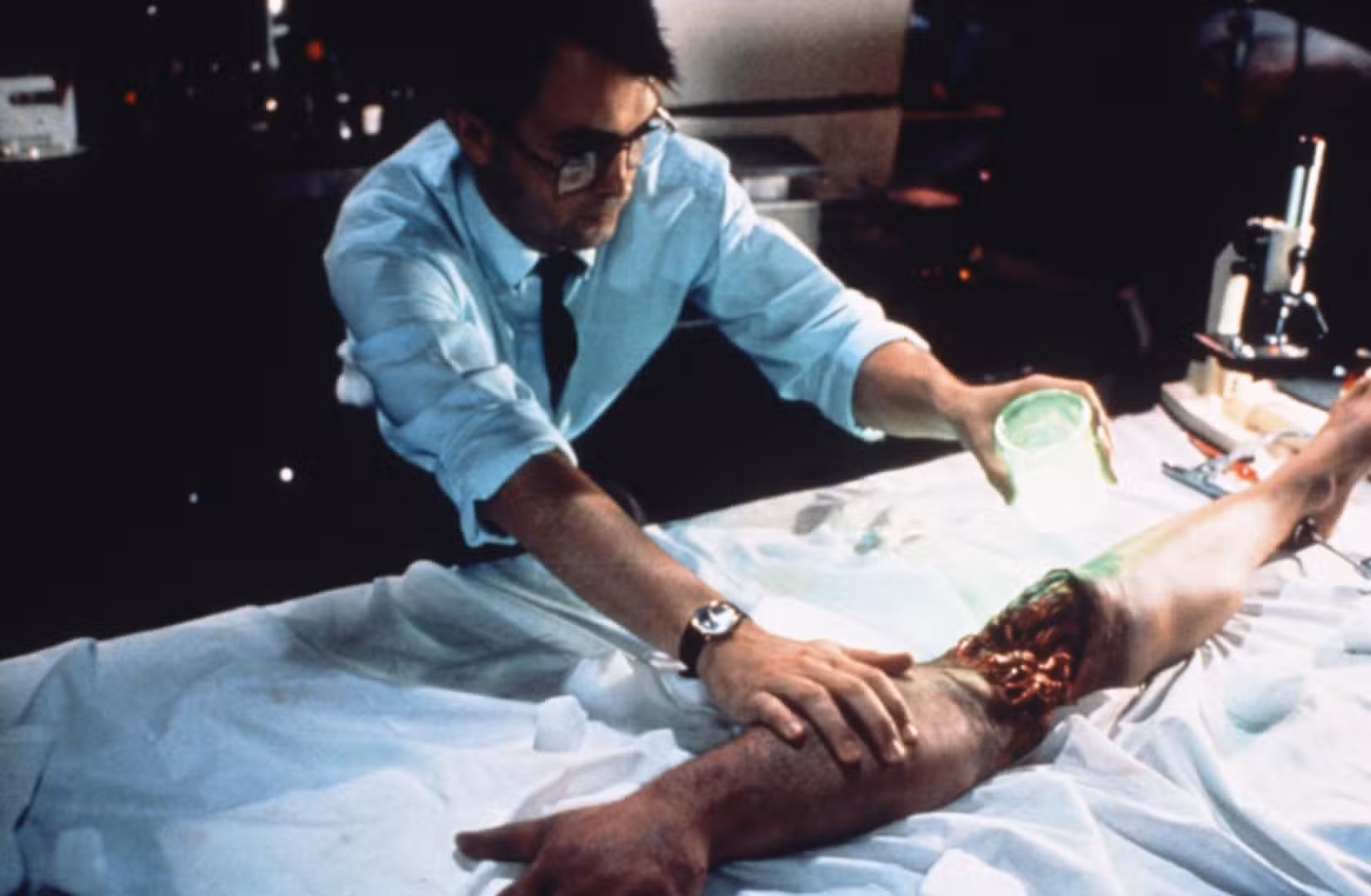 Um Herbert West de óculos usa seu reagente neon em um braço e uma perna decepados unidos em Bride of Re-Animator