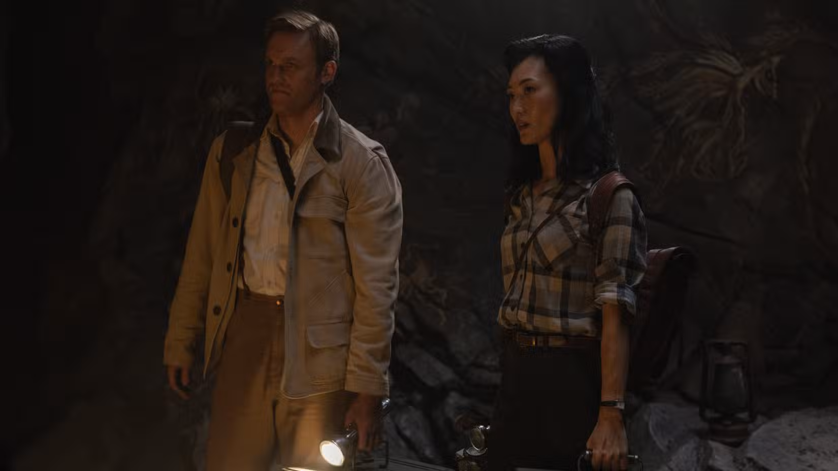 Lee Shaw (Wyatt Russell), um homem loiro segurando uma lanterna e parecendo chateado, fica ao lado da Dra. Keiko Miura (Mari Yamamoto), uma jovem que parece fascinada por algo em uma caverna em Monarch: Legacy of Monsters